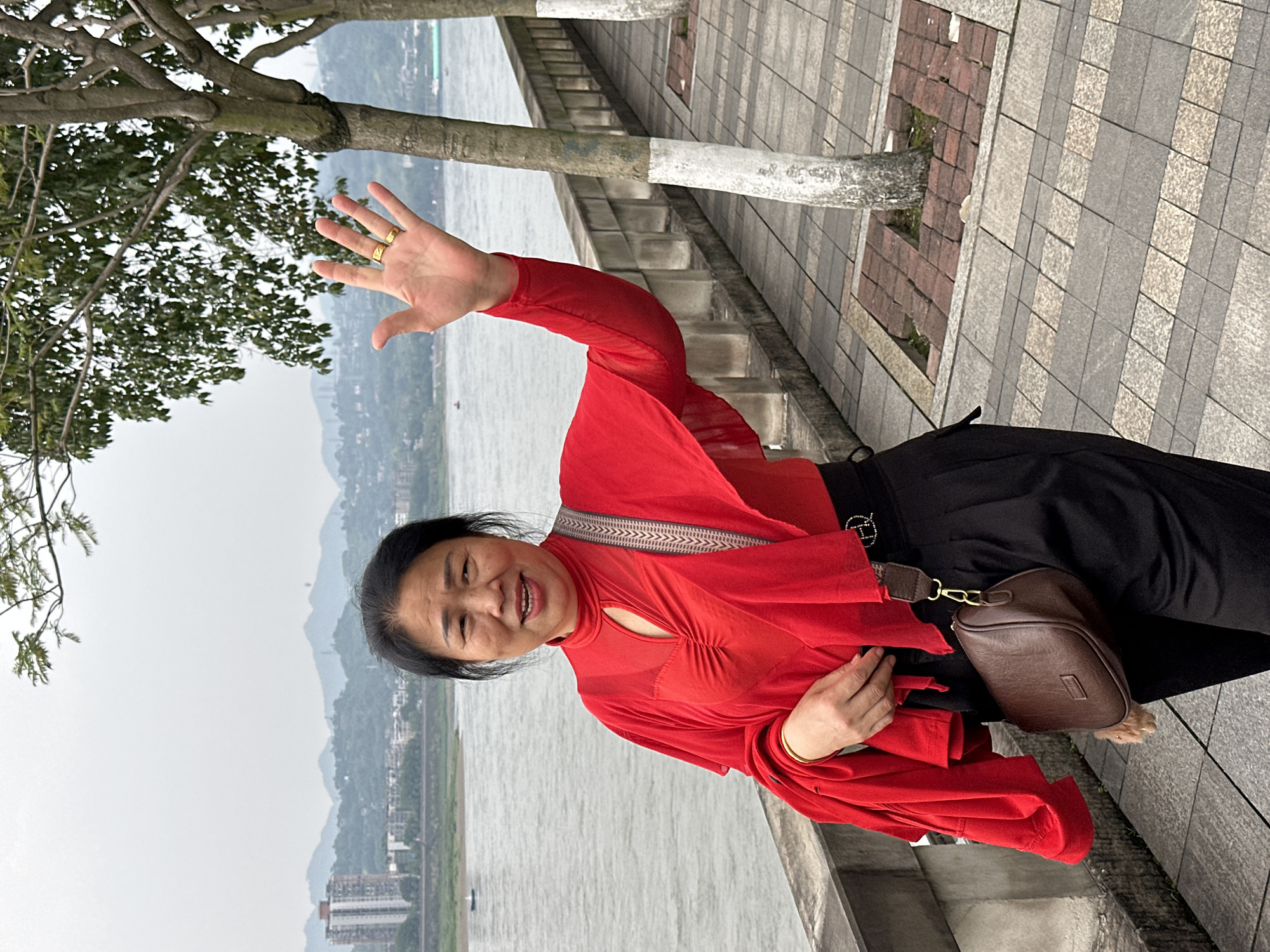 The image captures an adult woman in Jiangjin, China, positioned on a tiled walkway along a body of water. The water, reflecting a cloudy sky, is framed by a shoreline with visible buildings and distant hills. A tree with a portion of its trunk painted white stands to the left of the woman. She is wearing a bright red top, black pants, and a brown leather purse. Her right hand is raised in a wave, displaying rings on her fingers. The background suggests a location with both urban and natural elements.