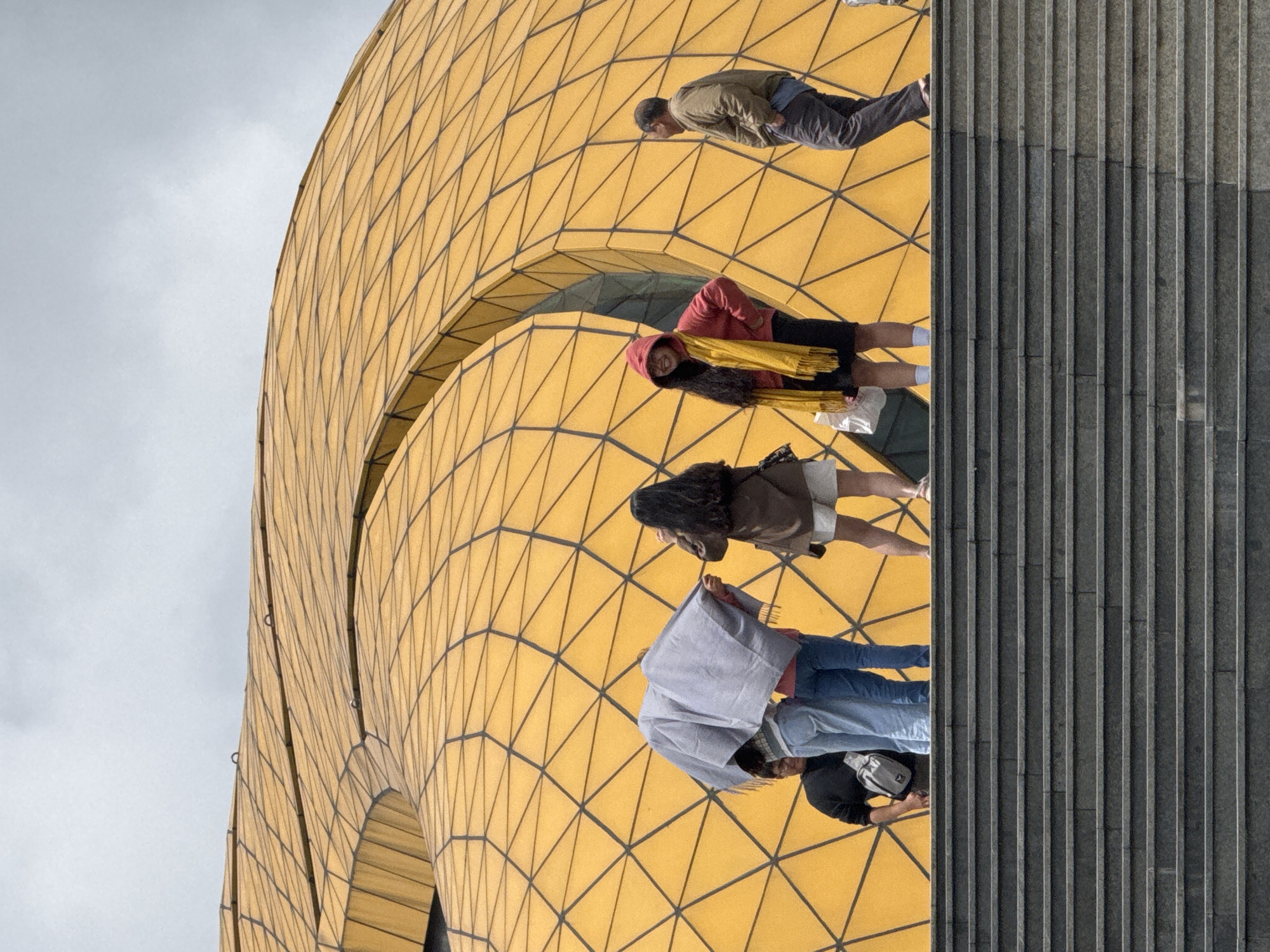The image shows a group of people standing on a set of outdoor stone stairs, in front of a large, undulating building with a distinctive yellow, diamond-patterned facade. The building's architecture is organic and fluid, resembling a giant, golden flower or shell. The sky is overcast, suggesting a cloudy day.
On the stairs, from left to right, are:
- A person wearing a black sweatshirt and a grey crossbody bag, partially obscured by a person in front.
- Two individuals standing together, one in light blue jeans and the other in darker blue jeans. They are mostly covered by a large, light grey draped fabric, possibly a shawl or blanket. The person on the right, wearing the jeans, is holding the fabric.
- A person wearing a brown jacket and white shorts, facing right and holding a camera or phone to their face, seemingly taking a photo.
- A person in a bright red hooded jacket and a yellow scarf, smiling and looking towards the viewer. They are wearing a black skirt and white socks.
- A man wearing a light brown jacket and dark pants, facing right with his hands in his pockets, looking away from the camera.
The yellow building's facade is composed of numerous triangular panels. The overall scene suggests a tourist attraction or a public gathering space. Based on the architectural style, this building is likely the Da Lat Flower Garden dome in Da Lat, Vietnam, known for its unique, petal-like structure.
