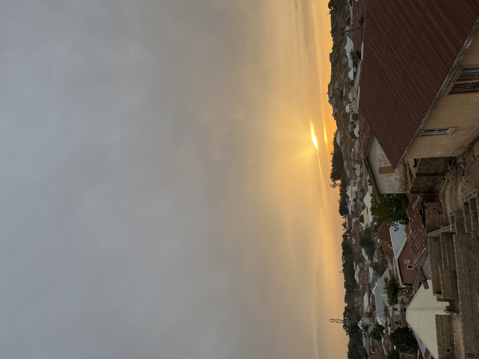 The image shows a landscape in Jos, Nigeria, at sunset. The foreground includes the roofs of various buildings and a building under construction with exposed brick. The middle ground consists of dense urban housing with visible roads and vegetation. In the background, a hazy horizon with a bright sun setting, illuminating the sky in shades of orange and yellow. The sky is partially obscured by clouds. A telecom tower is visible in the distance.