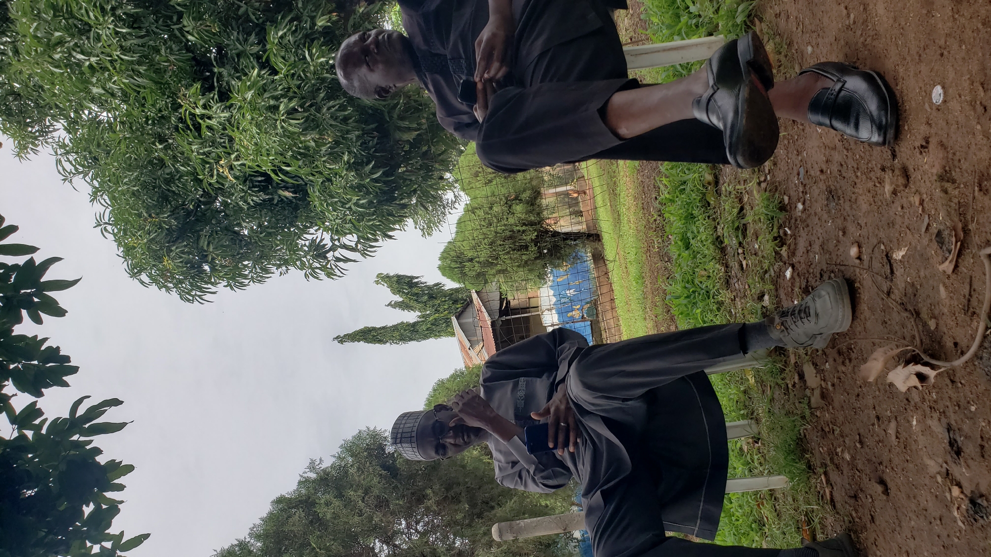 Two men are seated outdoors on white plastic chairs, facing towards the viewer, in what appears to be Unguwar Shanu, Nigeria. The man on the left is wearing a dark grey cap with a checkered pattern, glasses, and a matching dark grey traditional outfit. He is holding a smartphone in his right hand and gestures with his left hand. The man on the right is wearing a black traditional outfit and is looking to his right, holding a smartphone in his hands. Both men are wearing shoes; the man on the left has on grey sneakers, and the man on the right has on black loafers.
The background features lush green trees, some with large leaves, and a cloudy sky. A rustic building with a corrugated metal roof is visible behind a wire fence. The ground is a mix of dirt and sparse green grass. The overall atmosphere is casual and relaxed, suggesting an outdoor gathering or break.