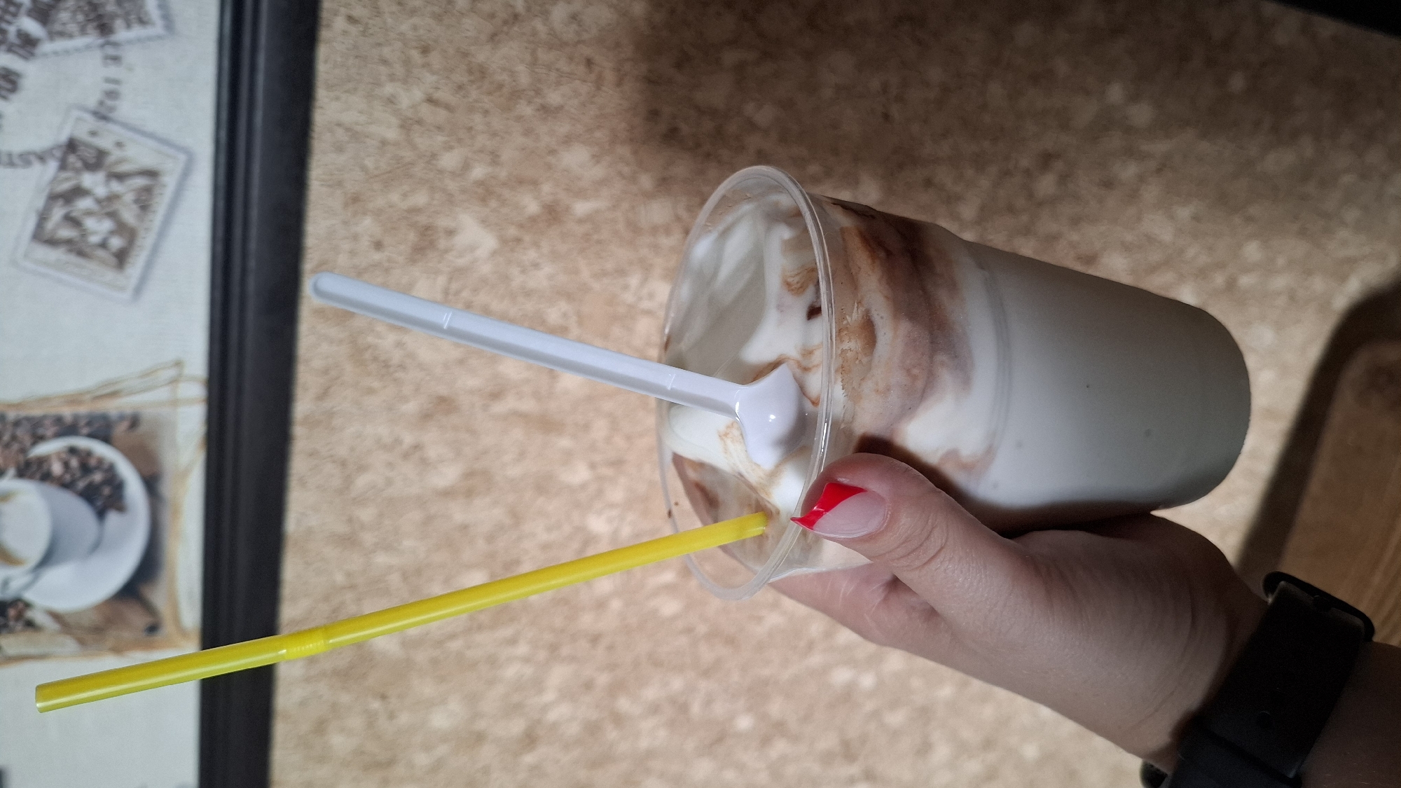 A close-up, waist-level, eye-level, vertical shot shows a hand holding a clear plastic cup filled with a dairy-based dessert, possibly a milkshake or soft-serve ice cream with chocolate syrup swirled throughout. A yellow straw and a white plastic spoon are inserted into the dessert. The hand is holding the cup from the right side, and the fingernails are painted with red tips and a French manicure. A black smartwatch is visible on the wrist. The background consists of a textured wall with a dark framed picture hanging on it, featuring what appears to be coffee beans and a cup of coffee. The lighting is neutral, and the overall scene is sharp and clear. There is no indication of the specific location within Shymkent, Kazakhstan.