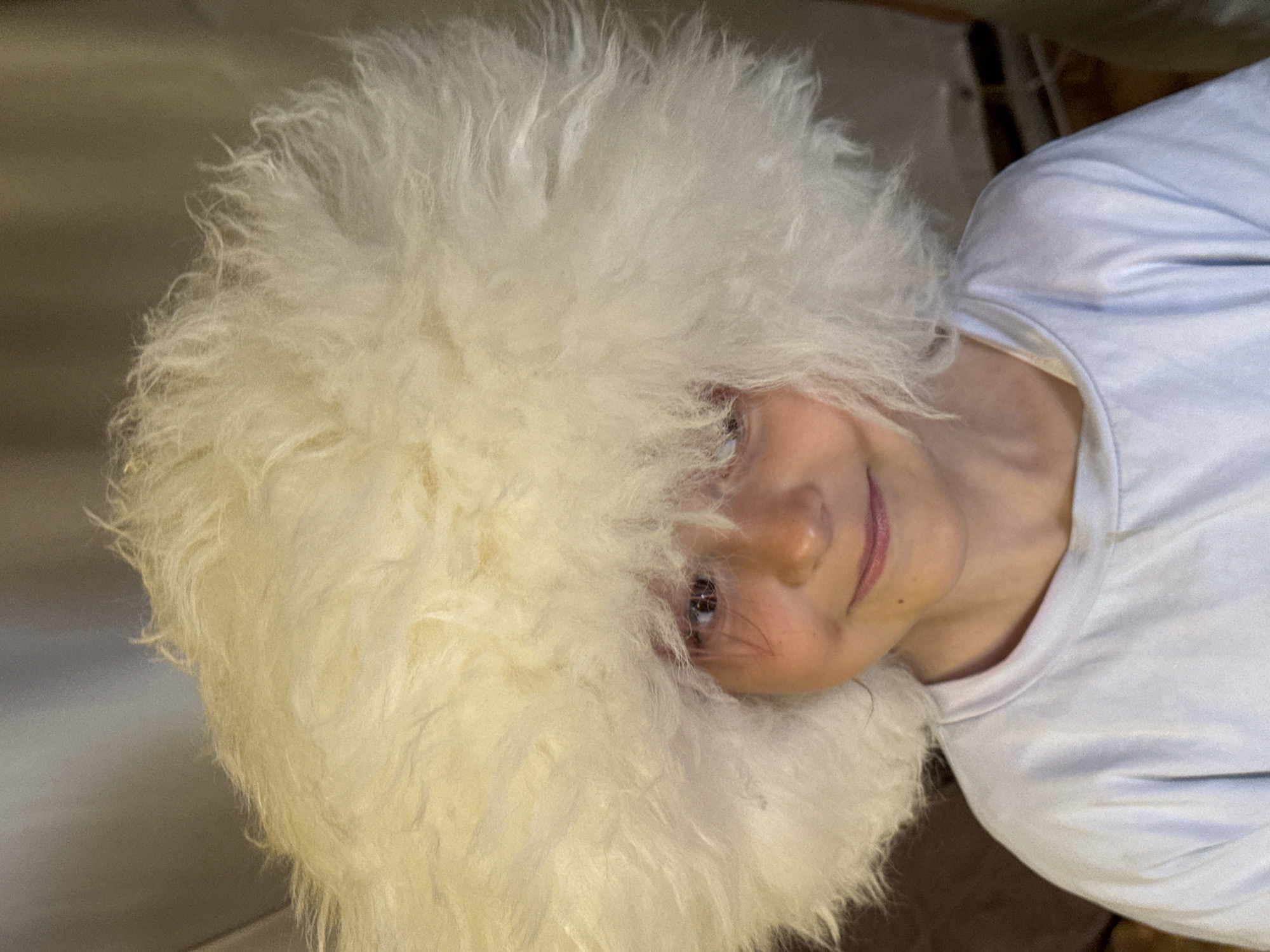 A close-up portrait features a young child, estimated to be between 5 and 10 years old, with light skin and visible dark eyes. The child is wearing an oversized, fluffy, white or cream-colored hat, possibly made of sheep's wool or synthetic material, which frames the face and extends significantly above and around the head. A light blue, crew-neck t-shirt is visible on the child's torso. The child looks directly at the camera with a slight, closed-mouth smile. The background is shallow in depth of field, displaying indistinct, muted brown and gray tones, characteristic of an indoor environment. The exact location is unknown, in the city of Unknown, Unknown.