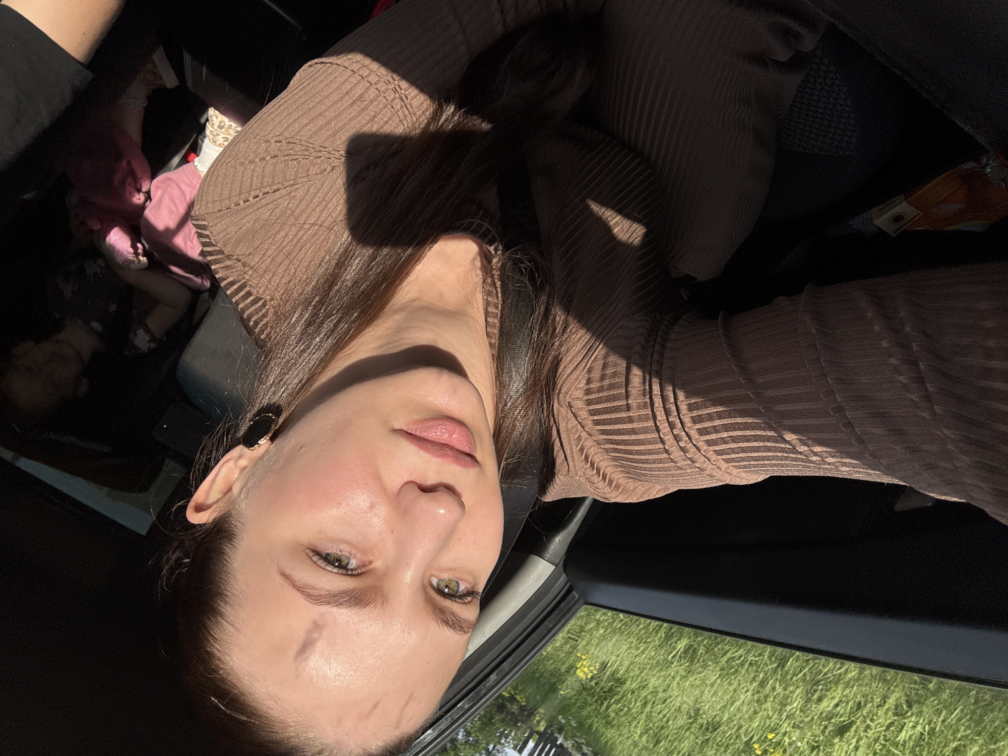 A young woman is captured in a selfie taken inside a car, likely in the front passenger seat, in Nizhnyaya Gar', Russia. She has long, dark hair, pulled back, and light green eyes. Her face is directly lit by strong sunlight, creating prominent shadows and highlighting minor marks on her forehead. She wears a brown, ribbed, long-sleeved top and a black oval earring with a gold rim on her left ear. A seatbelt strap crosses her chest.
In the background, to the left of the woman, a baby or toddler dressed in pink clothing is partially visible, possibly seated in a car seat. A leopard print pattern is visible on what appears to be a sleeve or sock near the child. The car's interior is dark, contrasting with the bright sunlight streaming in. Through the car window, a vibrant green field with scattered yellow wildflowers is visible under clear, sunny conditions, indicating daytime. There is no discernible text in the image.