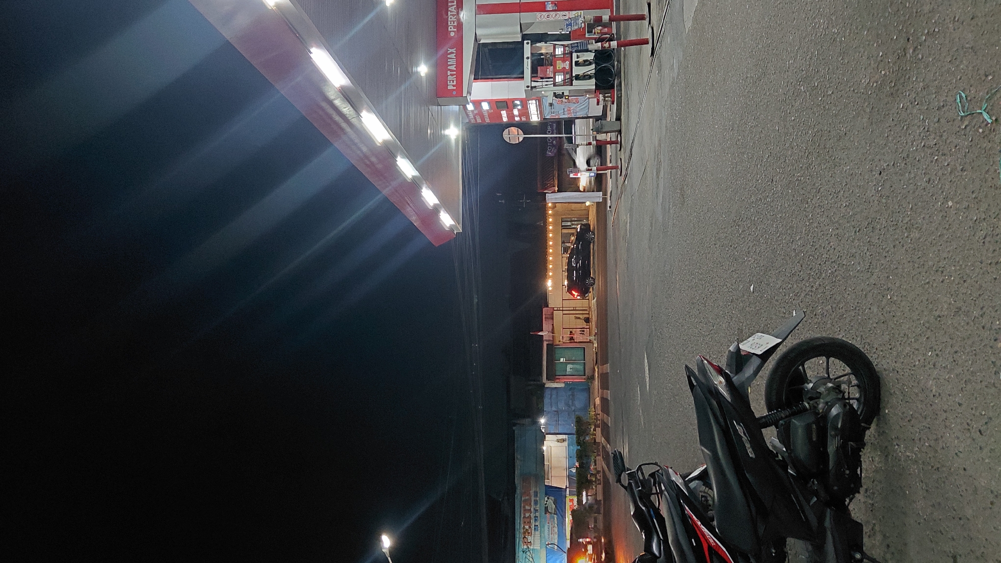 The image depicts a nighttime scene at a Pertamina gas station in Rejasari, Indonesia. In the foreground, a black motorcycle is parked on the asphalt. To the right, a gas pump is visible, with the Pertamax and Pertalite branding. Beyond the gas station, several buildings line the street. One building displays a sign with "FOTO COPY" and a red and white flag. A black SUV is parked in front of another building. In the background, the sky is dark, with the lights from the gas station canopy creating streaks of light. The overall environment suggests a functional, commercial area active at night.