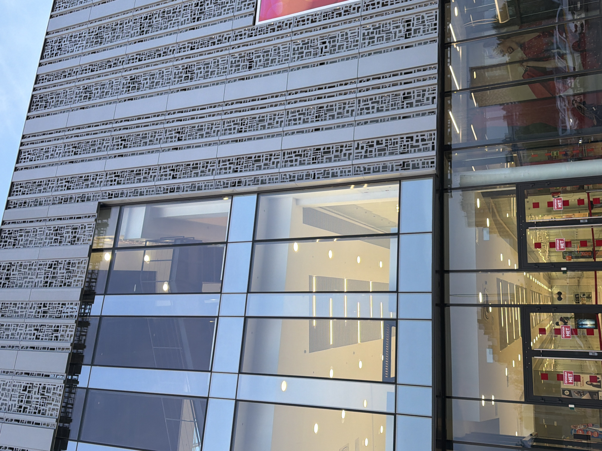 A low-angle, exterior shot captures the modern facade of a building, possibly a retail establishment, in an urban setting. The building features a large expanse of glass windows, reflecting the sky and surrounding structures. The upper portion of the facade is covered by a decorative screen with an intricate, geometric pattern. Red signs with the word "НЕТ" (meaning "No" in Russian) are visible on glass doors leading into the building, suggesting restricted access or a specific policy. Inside, the well-lit interior reveals what appears to be a shopping area with displays and merchandise. The overall impression is one of contemporary architecture with a functional interior design.