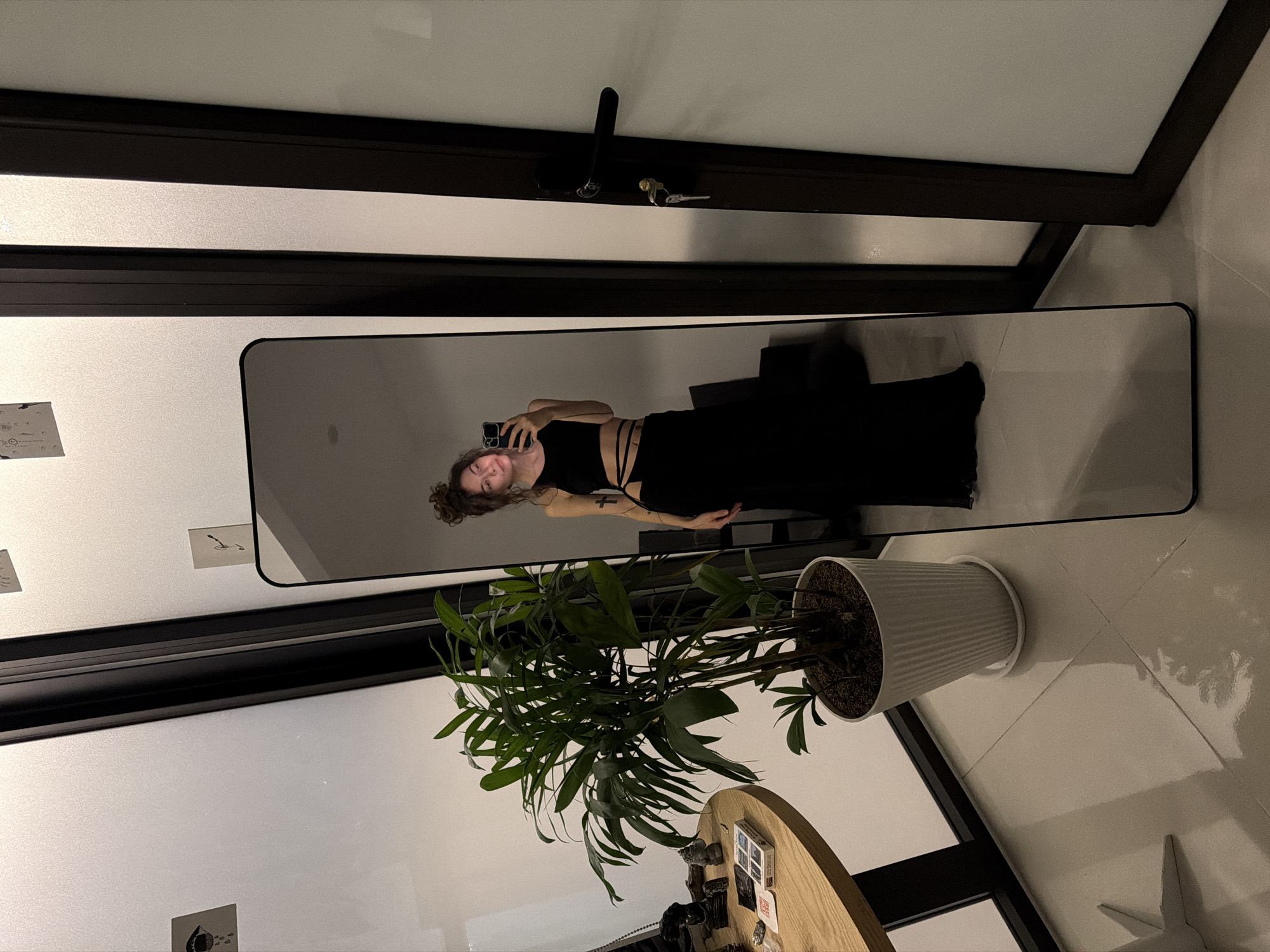 A mirrored reflection of a woman taking a selfie in Đà Nẵng, Vietnam. The woman has curly brown hair and a black outfit. Her reflection is in a floor-length mirror next to a potted plant and wooden table. The room is lit with ambient lighting. A door with a black frame is visible at the top. The floor is tiled. Artwork hangs on the wall. The table holds assorted items, including what appears to be playing cards and personal items.