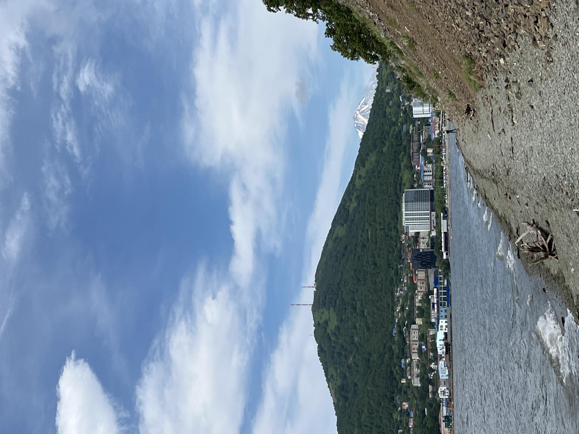 The image depicts a coastal scene in Petropavlovsk-Kamchatsky, Russia, with a body of water in the foreground and a city nestled at the base of a verdant, tree-covered hill in the mid-ground. A snow-capped volcano is visible in the distance, under a partly cloudy sky. The shoreline in the foreground is a mix of gravel and rocks, with some driftwood. A few small waves are lapping at the shore.
In the mid-ground, the city features a variety of buildings, including multi-story residential blocks and a modern, tall white building with many windows. Some smaller, colorful buildings are also visible. Two communication towers are situated on top of the hill. A single person is walking along the stony beach on the right side of the frame. The overall impression is of a city situated in a dramatic natural landscape.