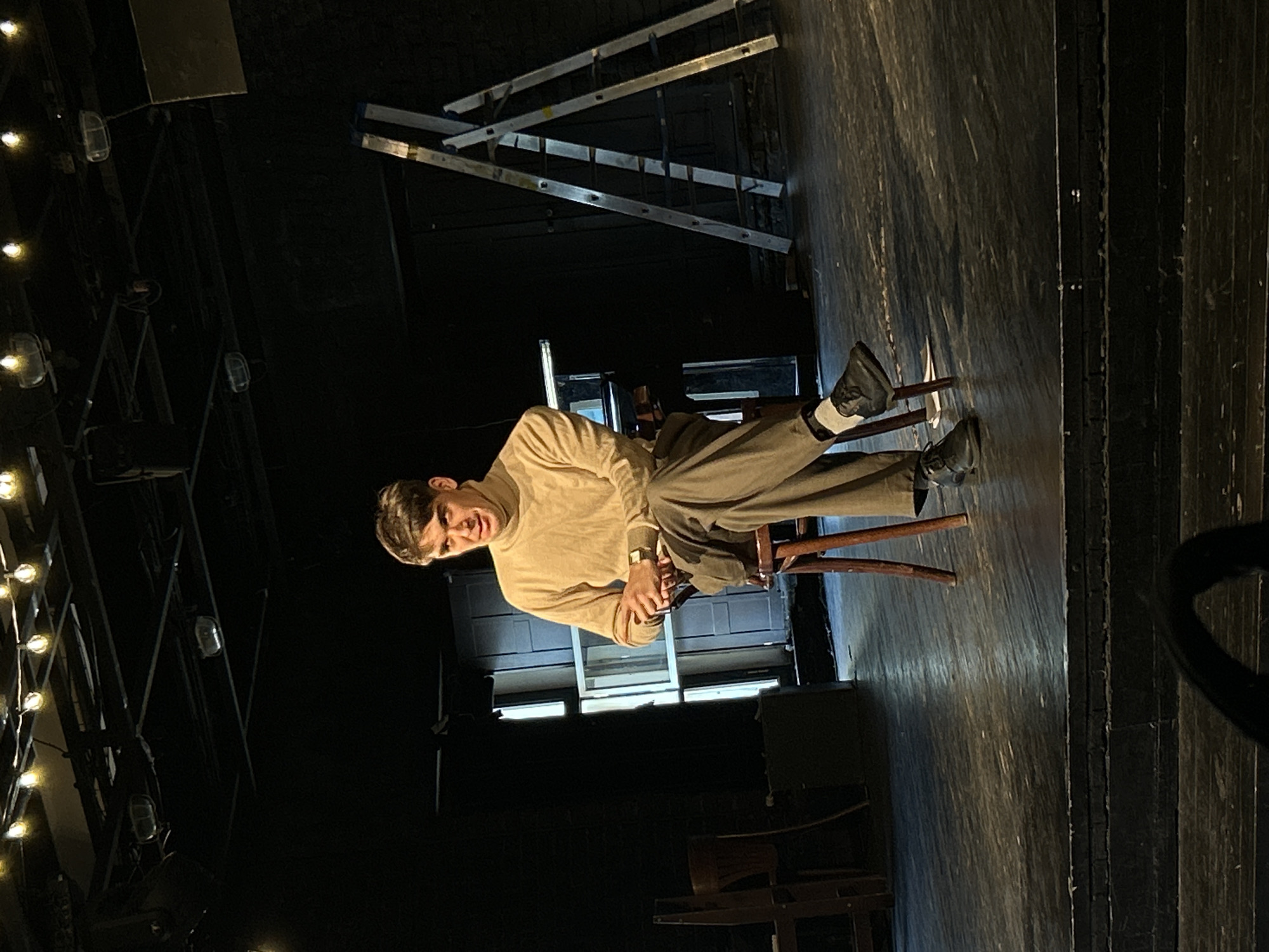 A young man sits on a dark wooden chair in what appears to be a dimly lit performance or rehearsal space. He looks directly at the viewer with a calm, slightly smirking expression. He is dressed in a light-colored turtleneck sweater, olive-grey trousers, dark shoes, and white socks, with a watch visible on his left wrist.
The floor beneath him is composed of dark, textured wooden planks. To his left, a tall structure holds multiple small, illuminated lights, resembling stage lighting or decorative string lights. In the upper background, two silver ladders are propped against a dark wall or backdrop. Behind the man, there is a rectangular structure with horizontal panes, possibly a window or a constructed set piece. The overall ambiance is dark, with focused lighting on the subject, suggesting an indoor setting like a theater or studio. There is no visible text in the image.