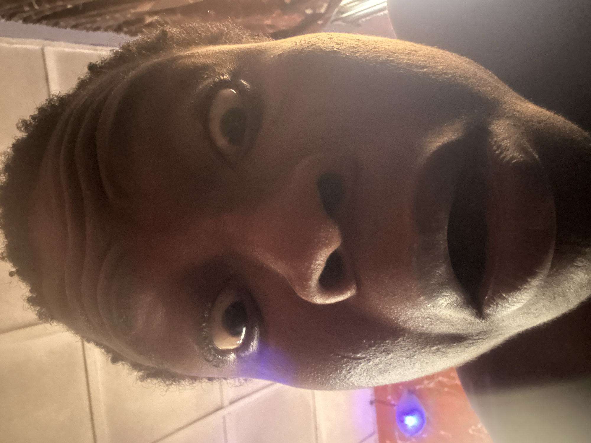 The image is a close-up, slightly rotated, selfie of a person with dark skin and short, curly hair. The person's face fills most of the frame. Their eyes are open and directed towards the camera. A portion of the mouth is visible and slightly open. The nose is centrally located. The background shows a white tiled wall on the left and a blurred, colorful light source near the bottom. The location and city are unknown based on the image.