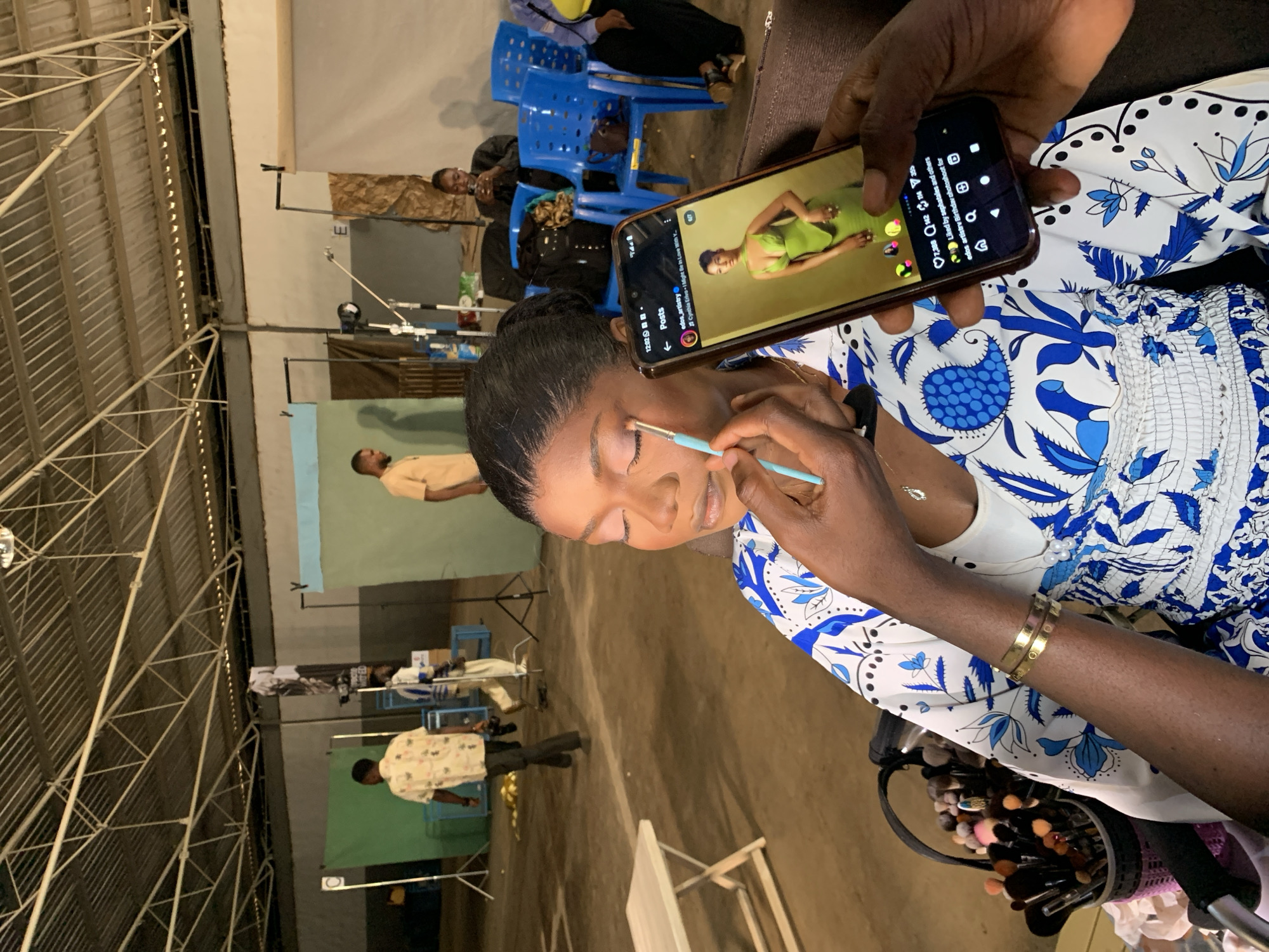 In Barakin Lerawi, Nigeria, a woman is having her makeup applied by a makeup artist in a studio setting. The makeup artist is using a light blue brush to apply eye shadow to the woman's closed eyelids. The woman is wearing a white top with blue floral patterns and a gold bracelet on her left wrist. A smartphone, held by another person not fully visible, displays a photo of a woman in a green dress. In the background, several people are visible, along with studio equipment like backdrops, lights, and chairs. The environment appears to be a large indoor space with a corrugated metal roof.