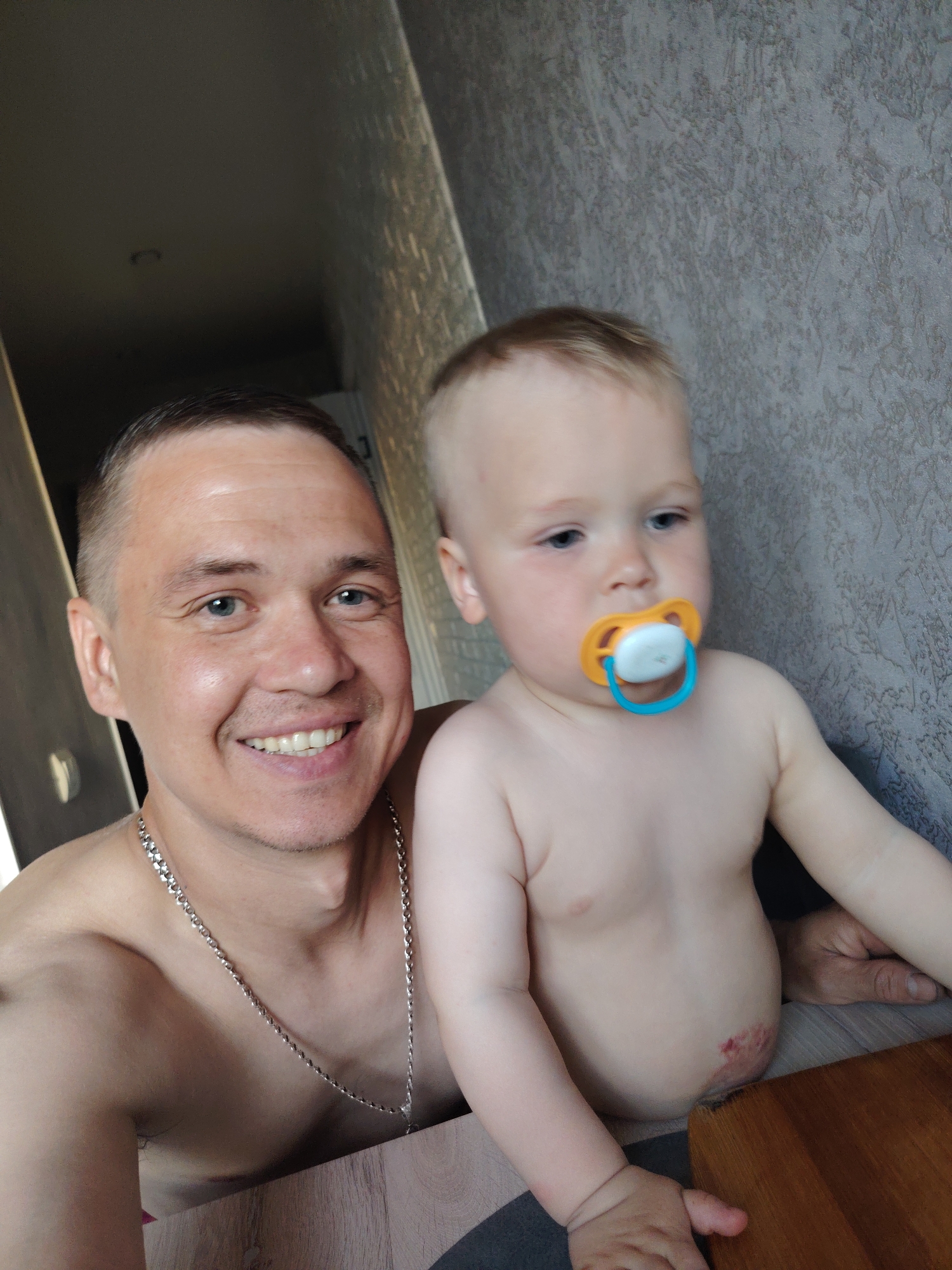 The image shows a man and a young child indoors. The man is fair-skinned with short brown hair, blue eyes, and a visible silver chain around his neck. He is shirtless and smiling. The child is also fair-skinned, with short blonde hair and blue eyes. The child has a pacifier in their mouth and is also shirtless. The background consists of a textured wall and a dimly lit doorway or hallway. The location is Chaikovsky, Russia. The man and child are likely interacting for a picture in a domestic setting.