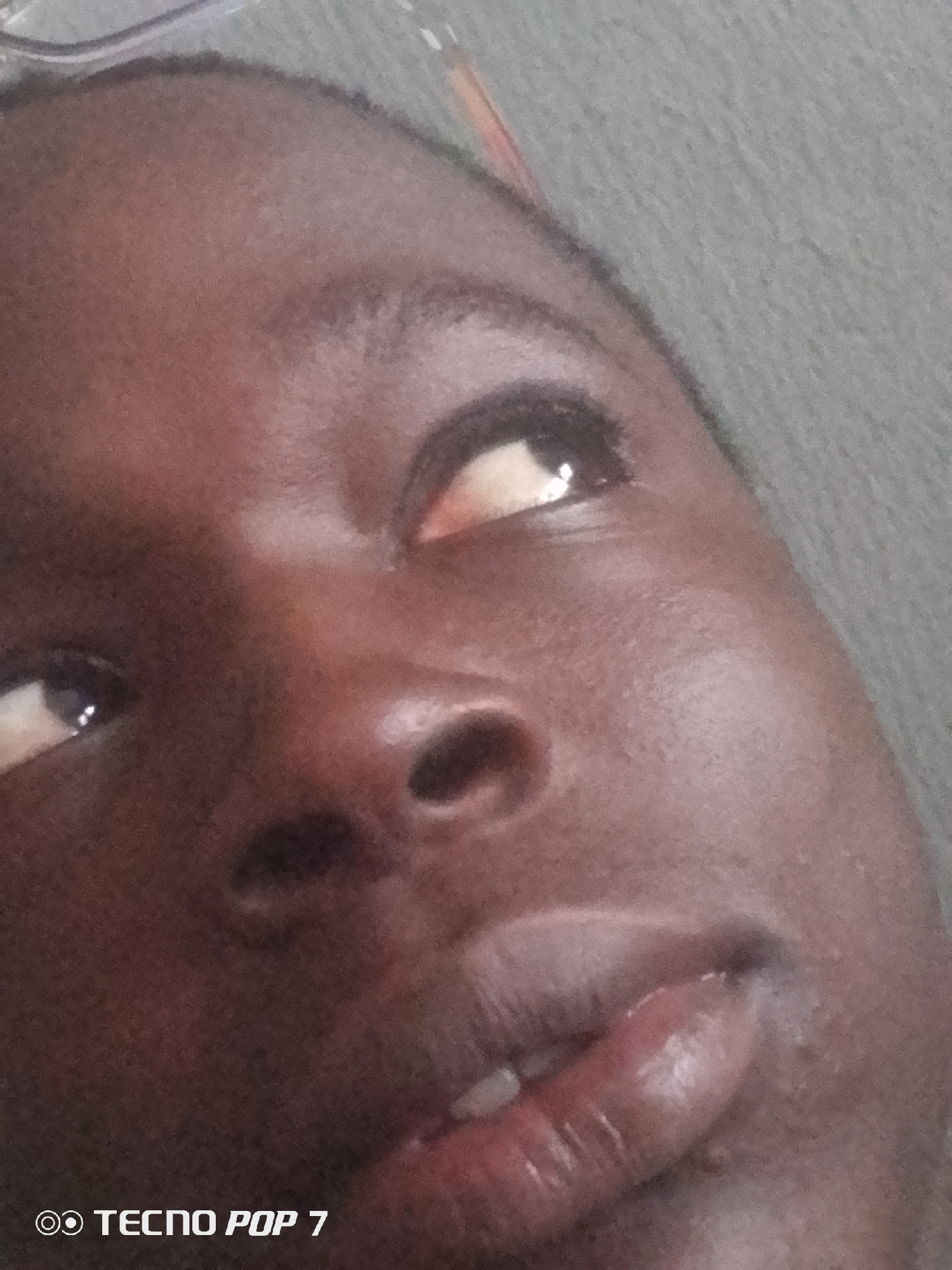 The image is a close-up, low-angle shot of a person's face, likely taken indoors given the artificial lighting and textured background. The subject has dark skin and is looking upwards and to the right of the frame. The focus is on the upper half of the face, prominently featuring the eyes and nose. The right eye is partially obscured by the angle, while the left eye is clearly visible with a bright reflection, suggesting the light source. The lips are slightly parted, revealing a glimpse of teeth. A watermark in the bottom left corner reads "TECNO POP 7". The setting appears to be a casual environment, possibly a home in Ipetumodu, Nigeria, but there are no specific environmental cues to confirm this. The overall mood is contemplative or perhaps a moment of awareness.