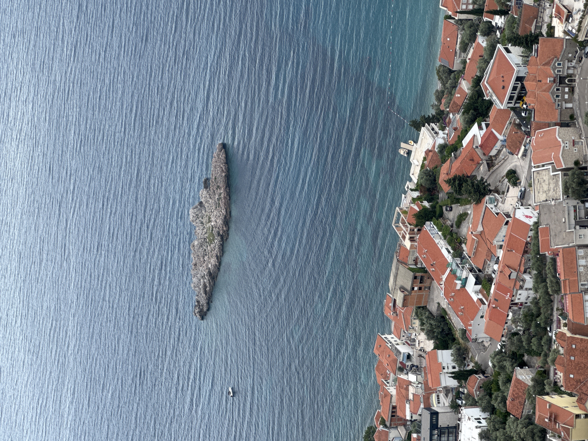 This aerial photograph captures a coastal town with a distinctive rocky islet situated just offshore. The scene is dominated by the vibrant blue of the sea, with rippling waves creating a textured surface. The rocky islet, a prominent feature, rises from the water, its rugged surface contrasting with the smoothness of the sea.
To the right of the islet and sea is a densely packed town. Buildings with terracotta-colored roofs are tightly clustered together, descending towards the water's edge. Lush green trees are interspersed among the structures, adding a touch of nature to the urban landscape. Streets wind through the town, with a few vehicles visible.
On the sea, near the islet, a small white object, possibly a boat or a buoy, is visible. Further out, a small boat is also discernible. The presence of these objects suggests human activity and perhaps leisure or transportation. The shoreline is marked by a thin white line, potentially indicating a path or a boundary.
The lighting suggests it is daytime, and the water's texture implies a slightly breezy condition. The overall impression is one of a picturesque coastal community, where human habitation harmoniously coexists with the natural beauty of the sea and its rocky formations. The location is Sveti Stefan, Montenegro. No visible text is present in the image.