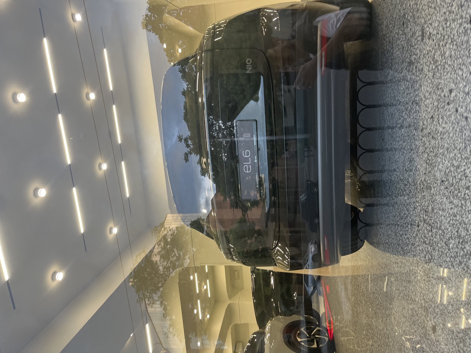 The image depicts the rear of a dark-colored NIO eL6 car parked in what appears to be a showroom in Yerevan, Armenia. The car is behind a glass window, and the reflection on the glass shows both the showroom interior (including ceiling lights and a curved wall) and exterior elements like trees and the sky. The car's license plate is visible, and the NIO logo is present on the rear. The vehicle is positioned on a rough, possibly concrete or pebbled surface. Other cars are visible inside the showroom.