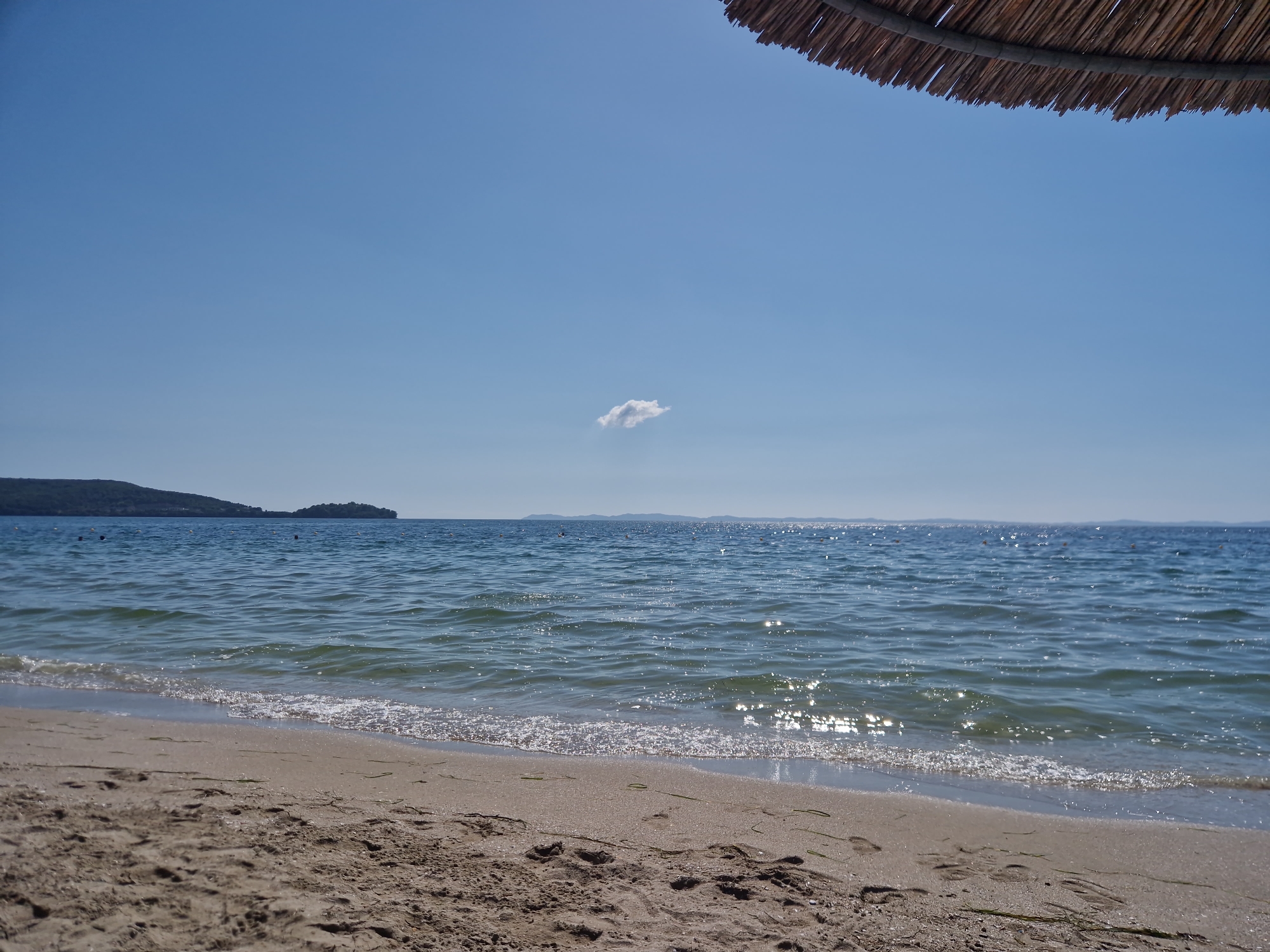The image depicts a serene beach scene in Igoumenitsa, Greece. The foreground is dominated by sand with visible footprints and scattered seaweed, leading towards the gentle waves of the Ionian Sea. The water is a clear blue, reflecting the bright sunlight and creating a shimmering effect. In the distance, a hilly coastline with vegetation lines the horizon. The sky is a vibrant, cloudless blue, with a single, small white cloud floating near the center. A portion of a thatched umbrella is visible in the upper right corner, suggesting a shaded area on the beach. The overall atmosphere is one of tranquility and natural beauty.
