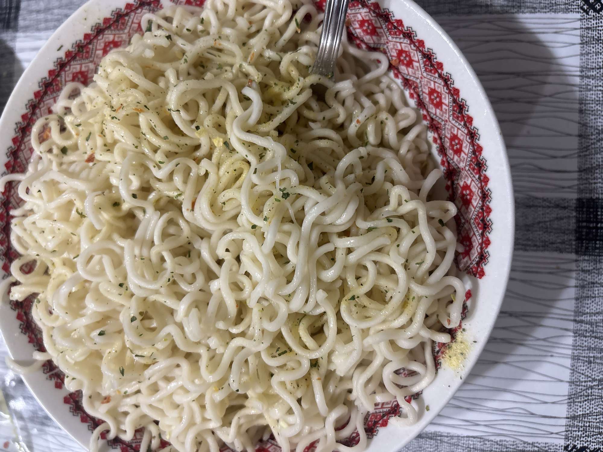 A close-up shot shows a white bowl filled with cooked noodles, topped with green herbs and a yellow powder. A fork is present in the bowl. The bowl has a decorative red and black pattern around the rim. The bowl sits on a grey and white checkered surface. The location is Pleven, Bulgaria.
