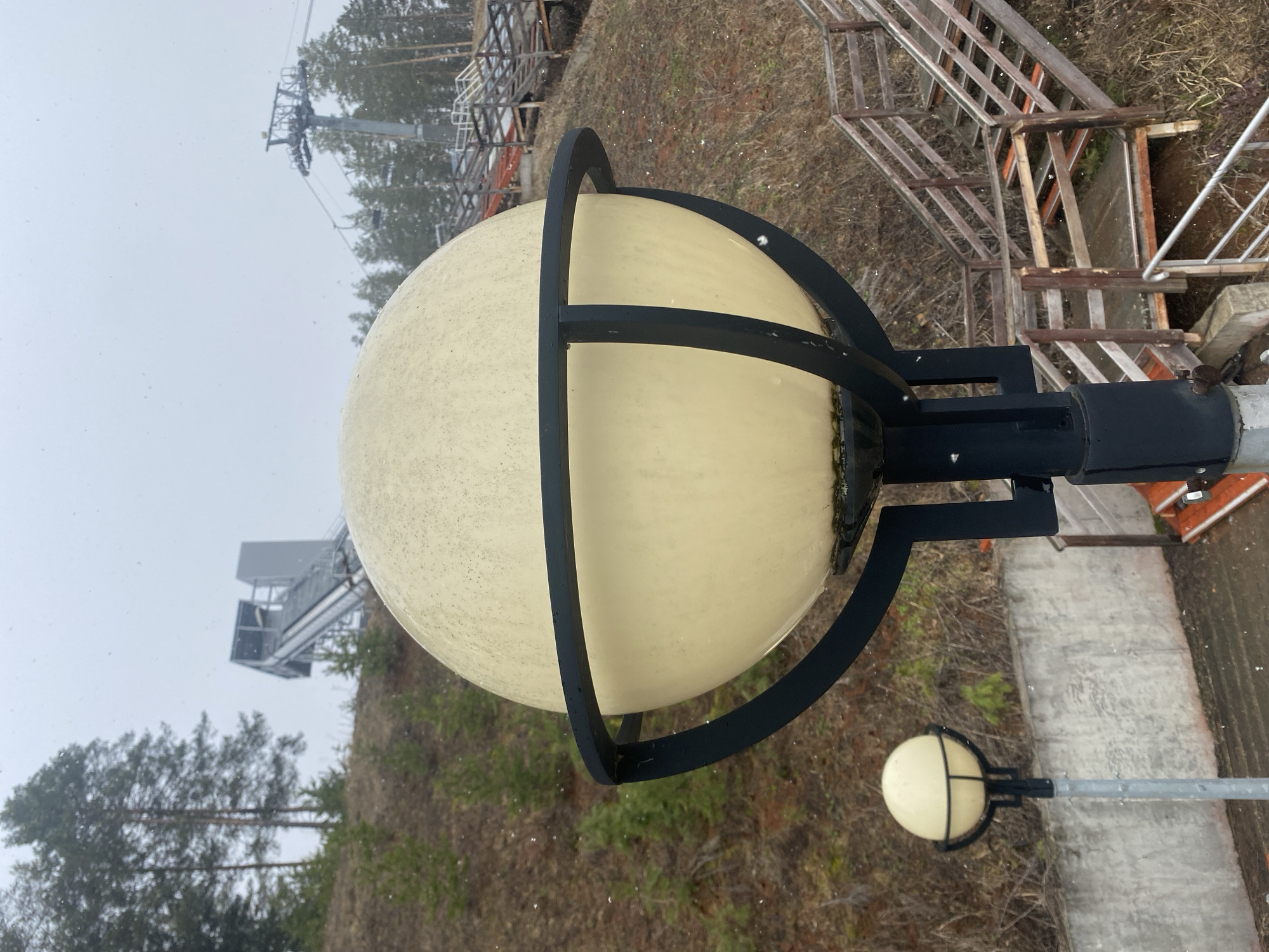 The image shows a close-up of a round, opaque lamp with a black metal frame, likely a street lamp or outdoor light fixture. It appears to be daytime with overcast lighting or snowfall. In the background, there's a blurred view of a ski lift or similar structure, along with trees and some wooden stairs. Another similar lamp is partially visible further down and to the right of the main lamp. The location is identified as Прикамский, Russia.