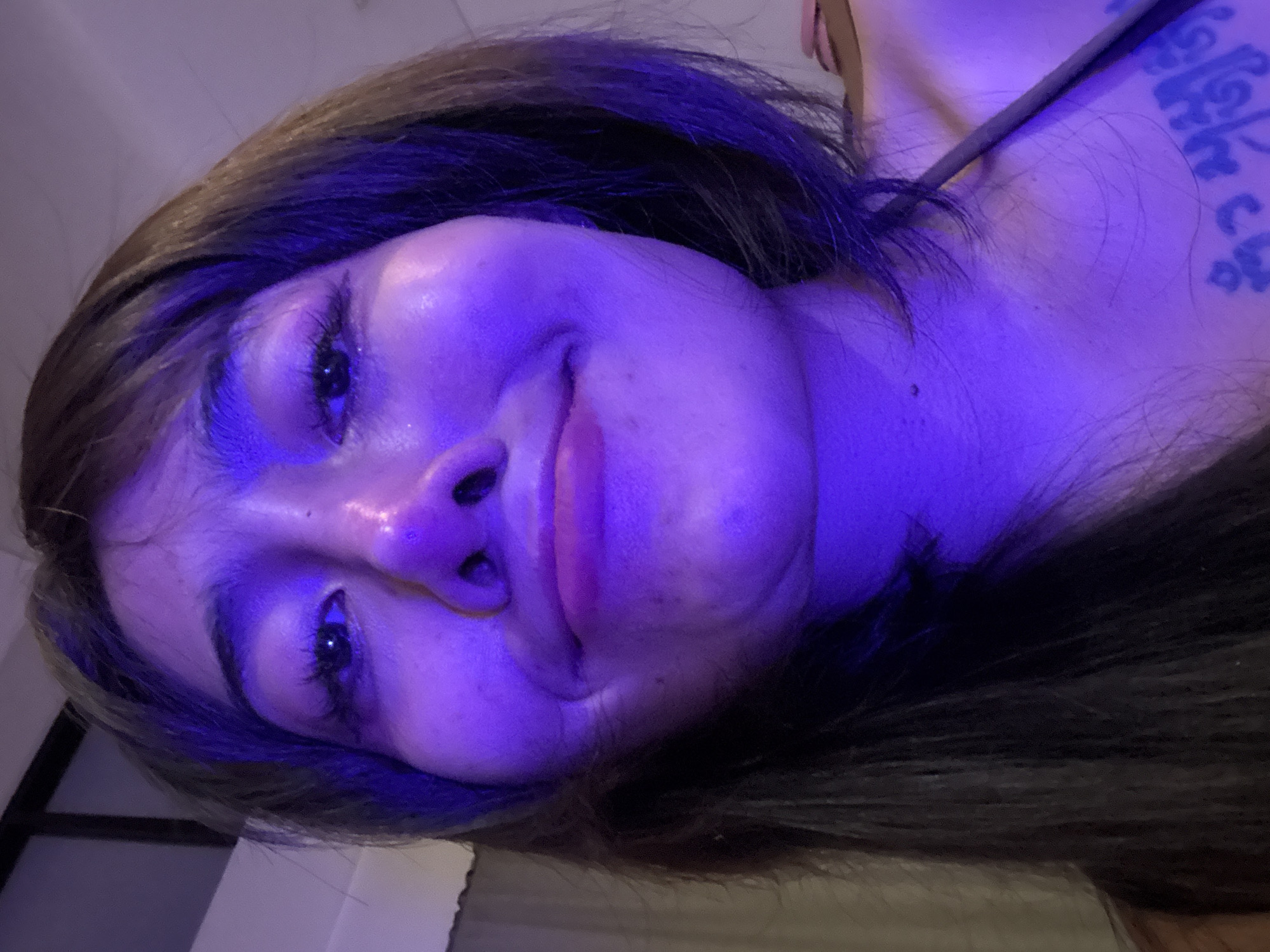 A close-up, tilted selfie of a person with dark hair and fair skin, illuminated by a strong blue light. The person is smiling slightly. A tattoo is visible on the right side of their upper chest/shoulder area. The background is indistinct, but it appears to be indoors with a window visible in the lower left corner. The location is unknown.