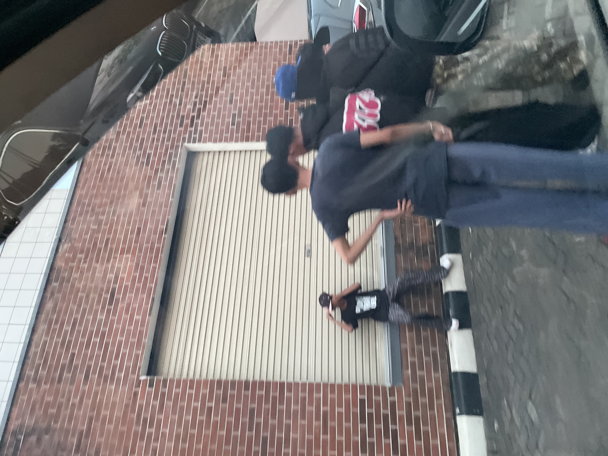 The image depicts a street scene in North Jakarta, Indonesia. A brick building with a large, closed, light-colored roller door forms a backdrop. Several people are present, some standing directly in front of the door and others further away. The individuals appear to be interacting, although the details of their actions are unclear due to the angle and distance. Cars are parked in the vicinity, and a black-and-white striped bollard is visible on the edge of a paved surface.