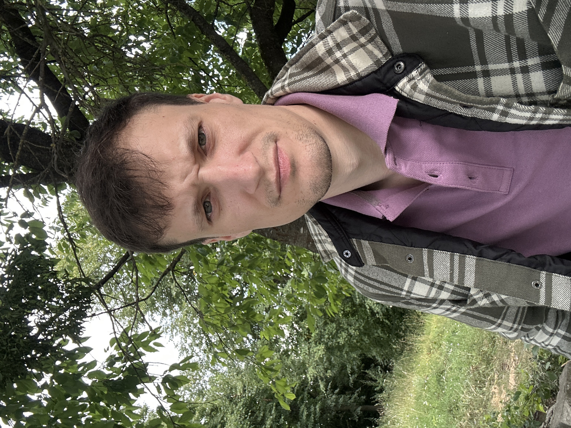A man is taking a selfie outdoors. He is wearing a purple polo shirt and a plaid jacket. He has short brown hair and is looking at the camera with a confused expression. The background is a park with trees and grass.