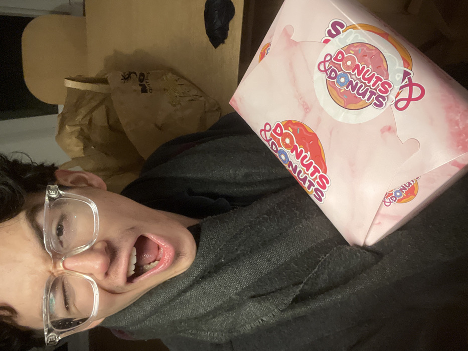 This media file captures a humorous indoor scene from Villeneuve-d'Ascq, France, featuring a person posing with a box of donuts. The main subject, a young person with dark hair and clear-framed glasses, is visible in the lower left, their face contorted in an exaggerated, wide-mouthed expression, conveying excitement or silliness. They are wearing a dark, possibly gray or black, hooded garment.
A prominent pink donut box from "Donuts & Donuts" is held in the upper right. The box features a light pink, marbled pattern and multiple logos displaying "DONUTS & DONUTS," often accompanied by a donut graphic.
In the background, behind the person and the donut box, there is a partially visible brown paper bag. Faint text, possibly "DON" or "NO ON" and a graphic resembling a spider, can be discerned on the bag. Further back, a larger, light brown object, which might be furniture or a cardboard box, is visible, along with a smaller black object.
The setting appears to be indoors, under artificial lighting, suggesting the photo was taken in the evening or at night. The individual's playful expression strongly indicates a mood of joy or anticipation, likely related to the donuts. The image itself is rotated 90 degrees clockwise.