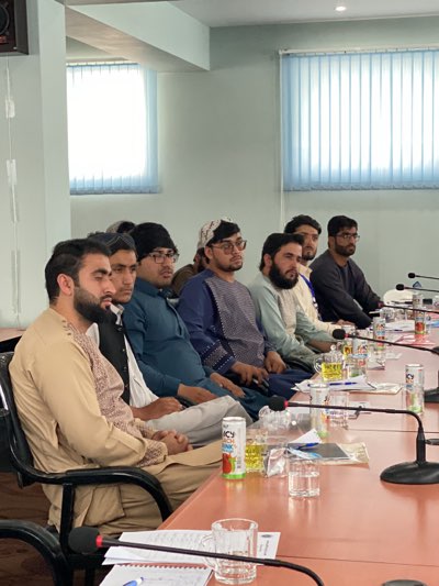 A group of eight men are seated at a long conference table in what appears to be a meeting room. The men are dressed in a mix of traditional and modern clothing. The table is covered with glasses of yellow liquid, a juice box, and microphone stands. The background features a window with partially closed blinds. This image was taken in Shurandam, Afghanistan.