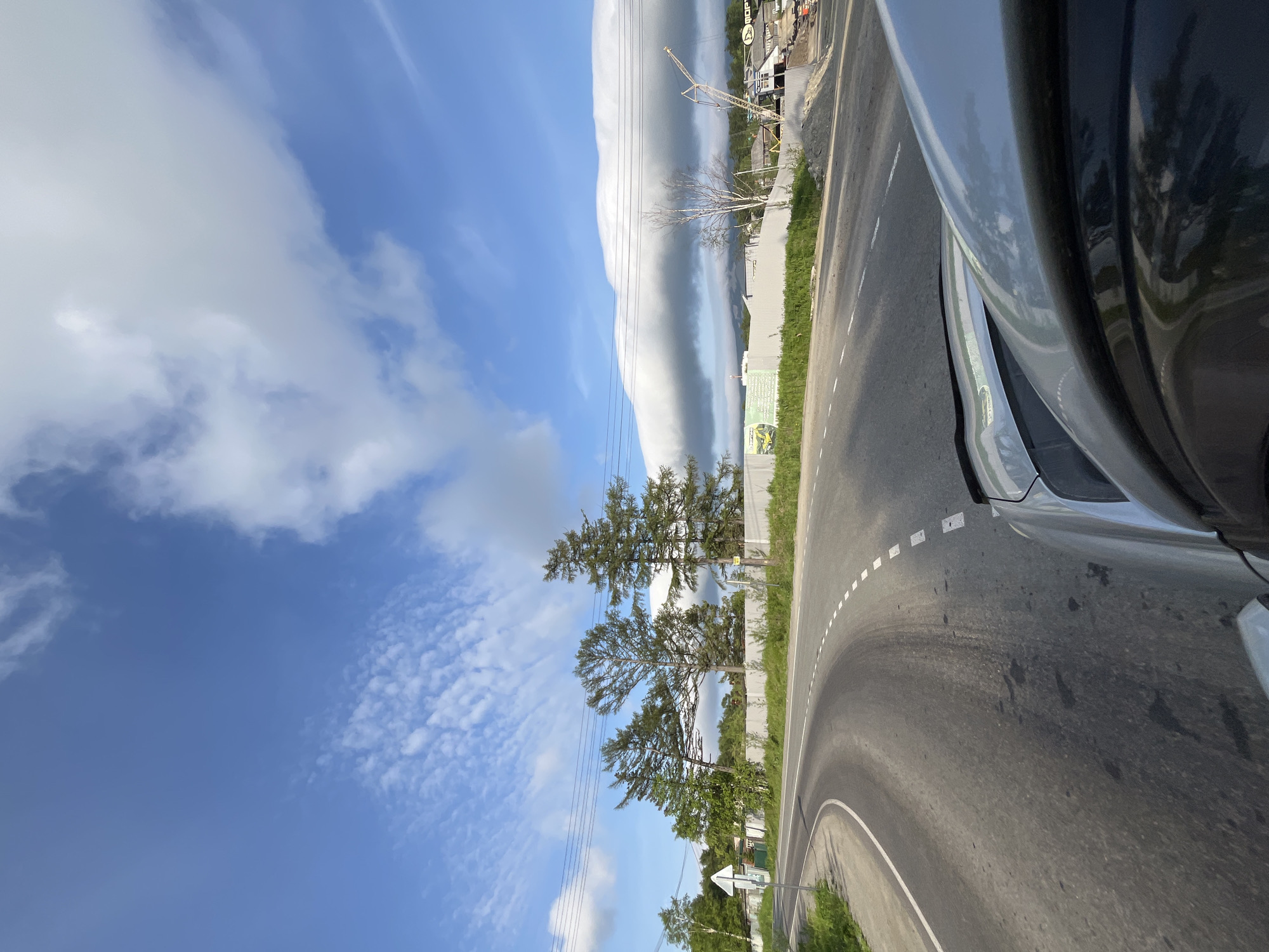 A fisheye lens view captures a bright, sunny day in Sovetskaya Gavan', Russia, as seen from the perspective of a vehicle traveling on a paved road. The road curves gently to the left, with white dashed lines marking the center and a solid white line on the left shoulder. Green grass and vegetation line the roadside, with a metal fence running along the right side of the road, behind which are trees and some buildings. The sky is a vibrant blue, adorned with scattered white clouds, including a dramatic, long, low-hanging cloud formation stretching across the horizon. Construction activity is visible on the right side of the road, with a crane and piles of sand or gravel. Power lines stretch across the top portion of the image. The car's silver hood and side mirror are visible in the foreground, reflecting the sky and trees.