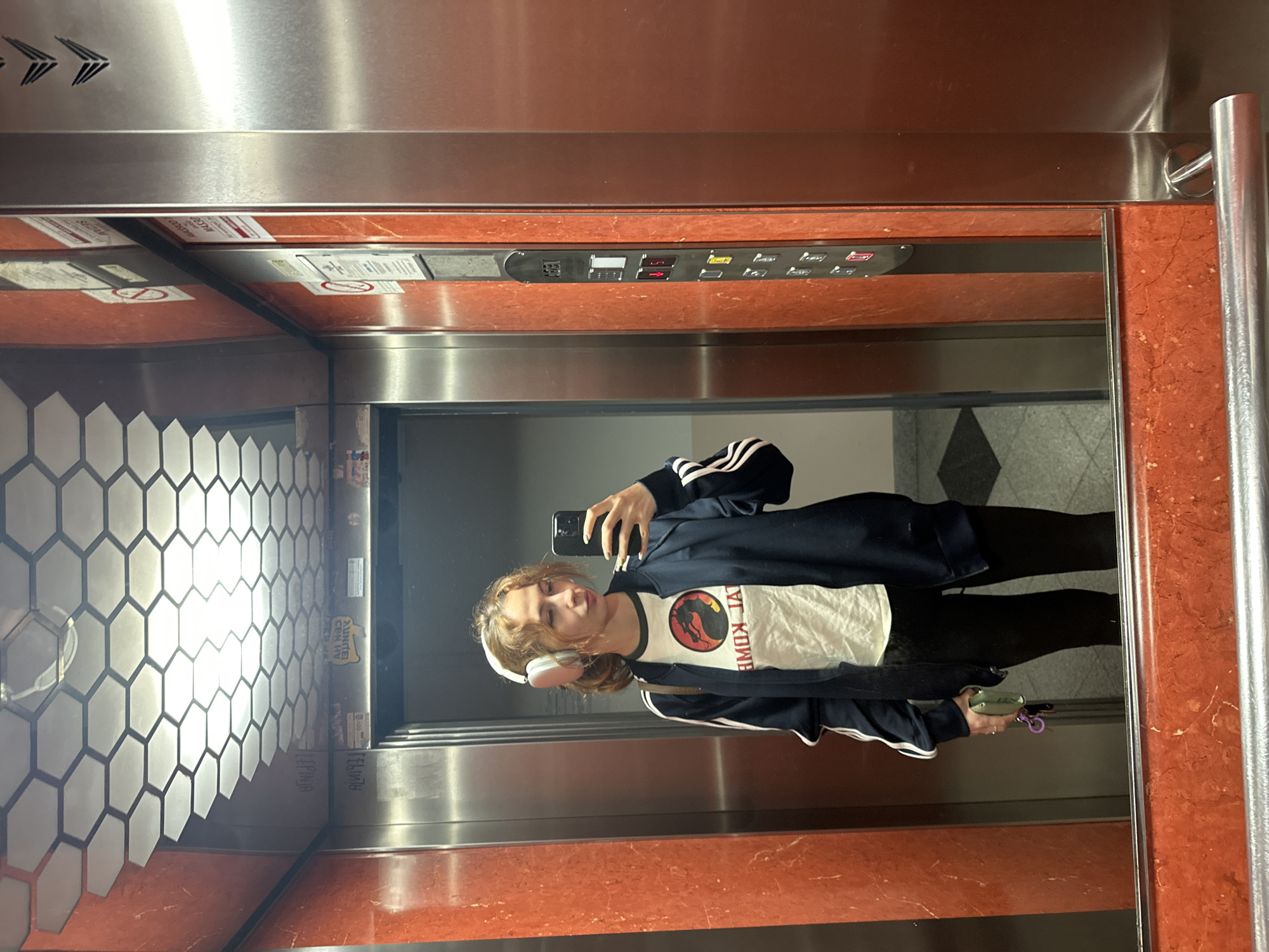 The image is a selfie taken by a person with medium-length brown hair, wearing white headphones, a black Adidas jacket with white stripes, and a white Jurassic Park t-shirt. They are standing in an elevator, holding a smartphone. The elevator has hexagonal tiles on one wall, a control panel with floor numbers, and a mirror that reflects the person and the interior of the elevator. The elevator's design incorporates orange-colored material. The location is Novi Sad, Serbia, as indicated by the advertisement on the elevator wall, which contains the word "JUNTER" and the words "CBH UV" (referring to a Novi Sad based company).