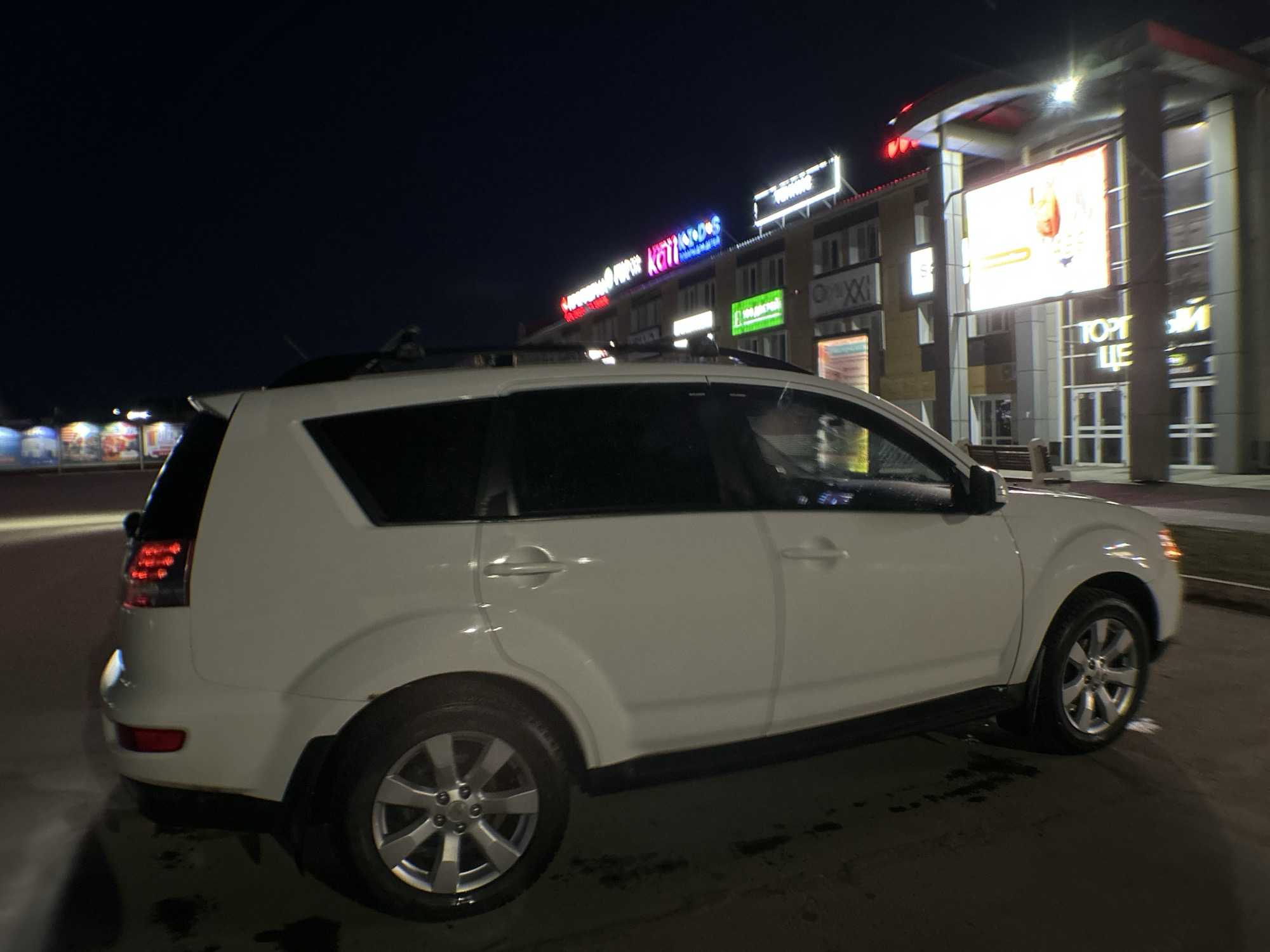The image shows a white SUV parked at night outside of the торговый центр «Универсам» in Chaikovsky, Russia. Multiple business signs are visible on the building. The scene is dark with artificial light from the building illuminating the car and surrounding area. No people are clearly visible.