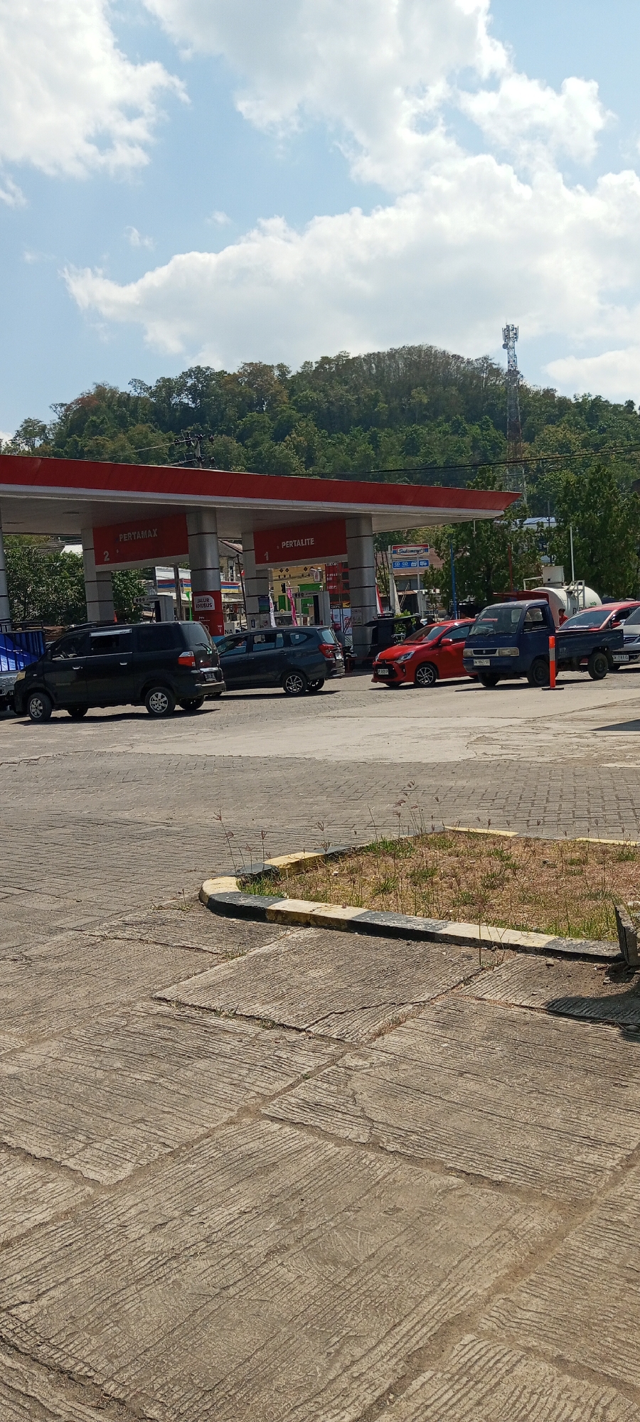 The image shows a gas station with several cars lined up to refuel. The gas station has a red roof with "PERTAMAX" and "PERTALITE" signs visible. The cars are parked on a concrete surface. In the background, there are trees and a hill with a communication tower on top. The sky is blue with some white clouds. The scene appears to be in a city in Indonesia, possibly Lempa.