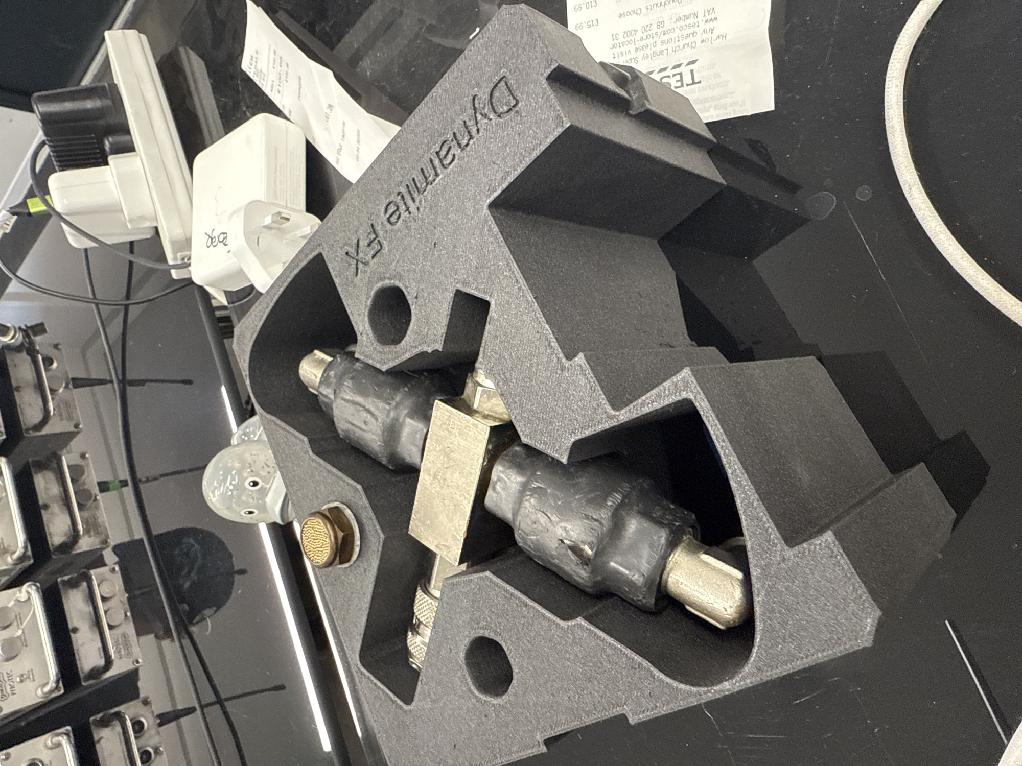 The image shows a black, foam-like object, labeled "Dynamite FX," encasing a metal fitting, possibly a valve or connector. The fitting has black coverings on either end. The foam piece is likely a custom-made container or organizer for the fitting.
In the background, various objects are visible, including electrical adapters, what appears to be a receipt from Tesco, and an array of metal components. The receipt indicates that Tesco is located in Harlow Church Langley Superstore. Given that Harlow is a town within the Epping Forest district of Essex, United Kingdom, the image could be taken in this general locale.