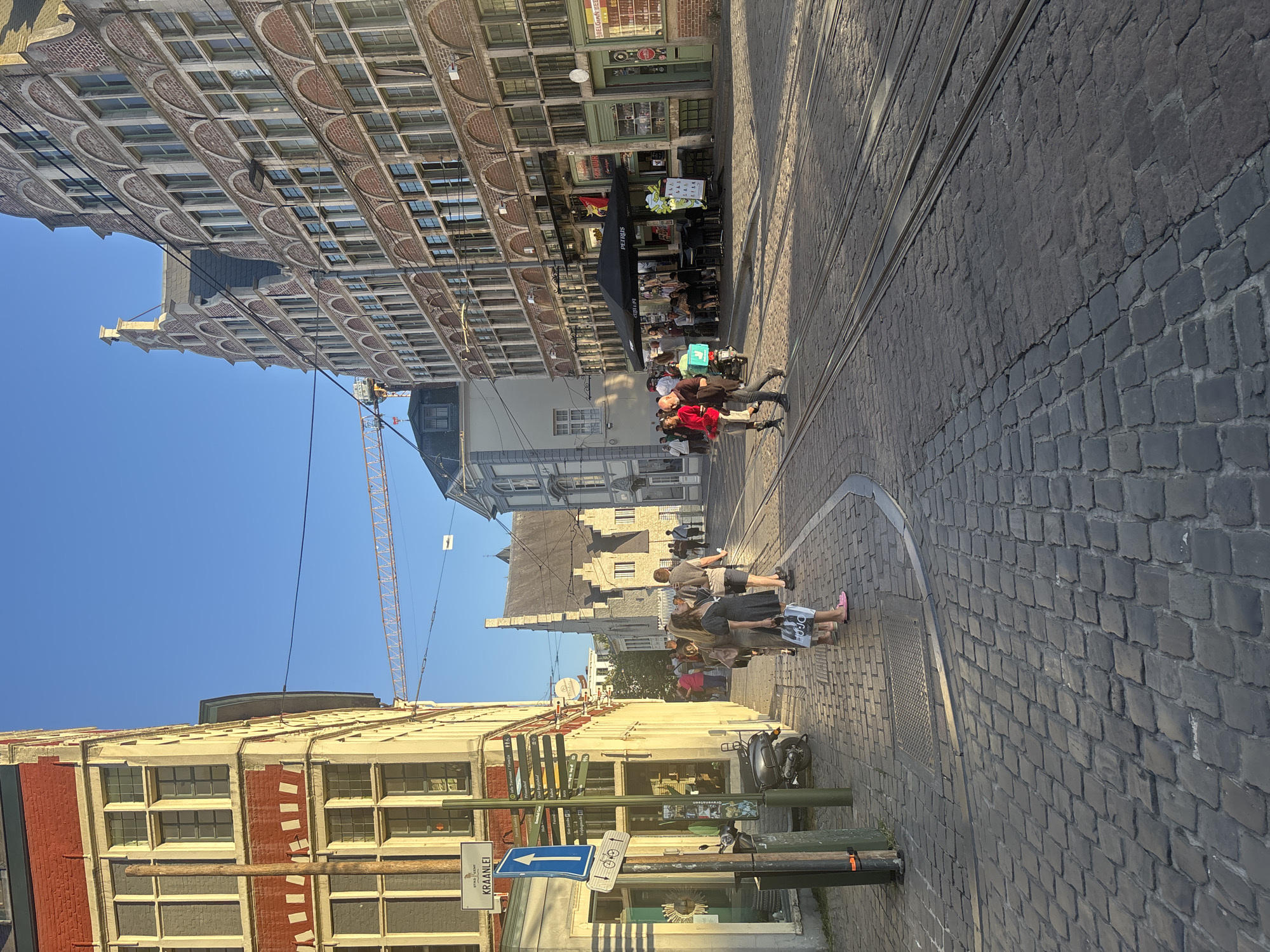 The image captures a view of the Kraanlei street in Ghent, Belgium, during a sunny day. The scene is dominated by historic brick buildings with characteristic Dutch gable roofs, lining a cobblestone street. Tram tracks are visible on the ground, indicating active public transportation.
On the left side of the street, a red and white brick building features large windows and a signpost with directional arrows, one pointing to "KRAANLEI." A scooter is parked next to the signpost. Several people are walking on the sidewalk and crossing the street.
In the middle of the street, a group of people are walking towards the viewer, some dressed in casual clothing. Further down the street, an outdoor seating area with umbrellas is set up in front of a building, suggesting a cafe or restaurant. A person in a bright yellow safety vest is standing near this area.
The sky is clear blue, and the sunlight casts long shadows, indicating it's either late afternoon or early morning. The overall atmosphere is that of a lively European city street with a blend of historical architecture and modern activity.