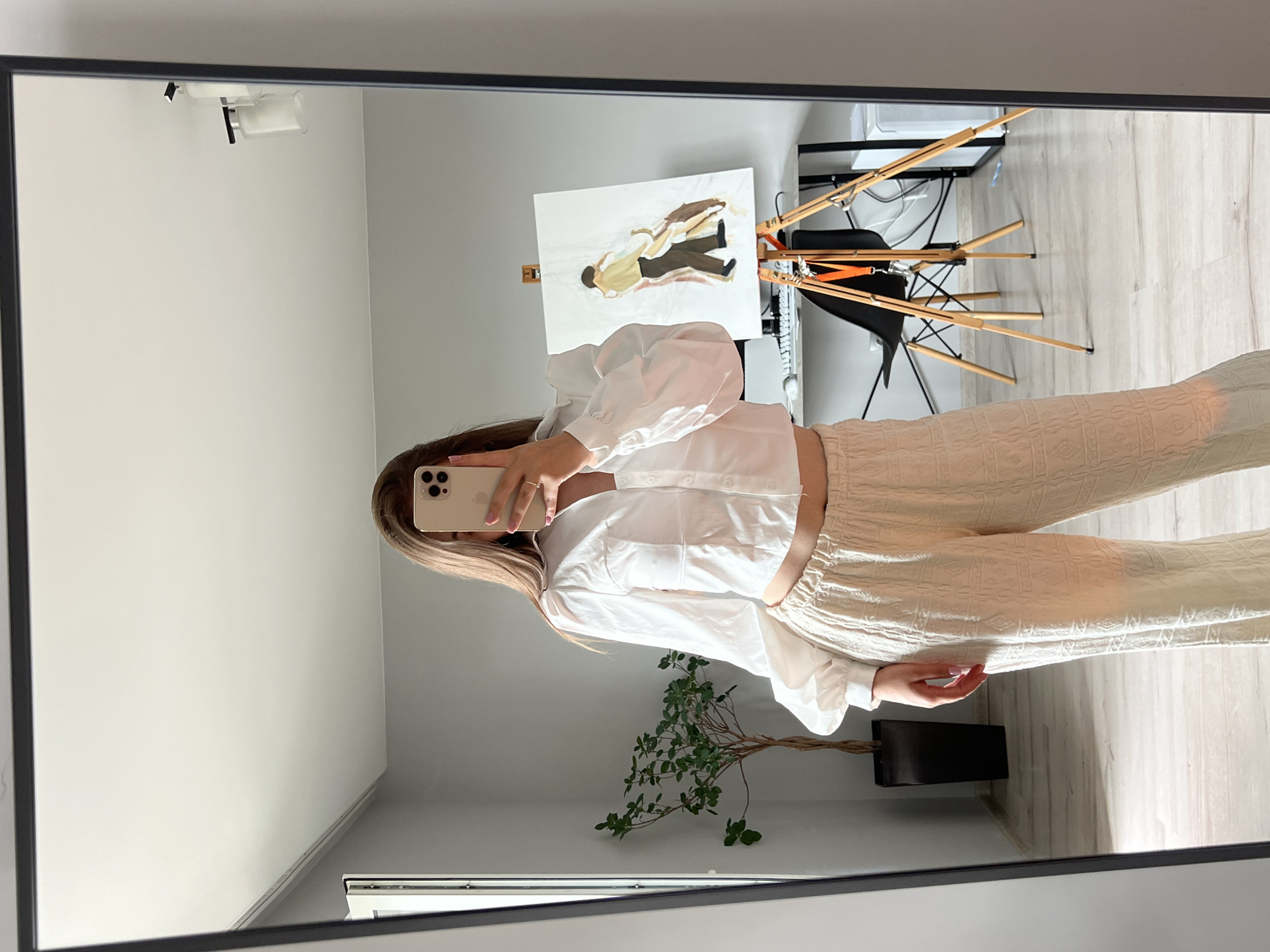 A person is taking a mirror selfie in a room. The person is wearing a white puff-sleeved crop top and loose, textured cream-colored pants. In the reflection, a plant in a black pot is visible on the left. Behind the person, there is a desk with a computer monitor, a black chair, and an easel with a painting of two figures. The room has light grey walls and a light-colored floor. The image is a photograph taken with a smartphone. There is no information available about the specific location in Ternopil, Ukraine.