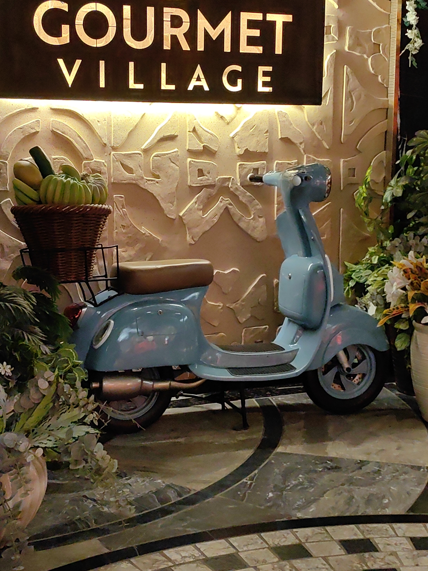 A light blue vintage scooter is parked on a patterned floor in front of a textured wall. Above the wall, a dark sign with illuminated gold lettering reads "GOURMET VILLAGE." A wicker basket filled with green gourds and a zucchini sits on the back of the scooter. To the left and right of the scooter are arrangements of green foliage and some white flowers. The scooter is facing to the right, with its kickstand down. The flooring is made of dark and light marbled tiles. The scene appears to be an outdoor or semi-outdoor decorative display. The text "GOURMET VILLAGE" suggests this may be the entrance to a restaurant or a food-related establishment.
