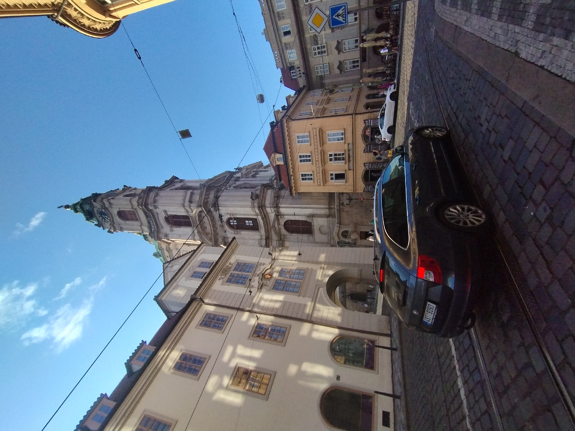 The image depicts a street scene in Prague, Czechia, with a notable architectural landmark in the background. The most prominent structure is a tall, ornate tower with a green spire, likely part of a church or historical building. This tower has a clock face visible. Adjacent to it are other buildings with cream and yellow facades, featuring multiple windows and red-tiled roofs. The street itself is paved with cobblestones and has tram tracks running through it. A dark-colored station wagon is parked on the street, facing away from the camera, and its rear brake lights are illuminated. A white car is visible further down the street. Several individuals are present, some walking and others appearing to be seated at outdoor cafe tables. Traffic signs, including a pedestrian crossing sign, are also visible. The sky is clear and blue with a few wispy clouds. The overall impression is of a sunny day in a historic European city. Based on the architectural style and the presence of the St. Nicholas Church tower, the location is likely Malá Strana Square in Prague.