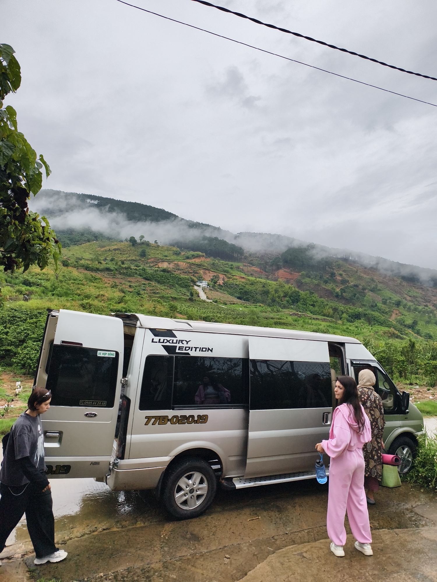 The photograph depicts three individuals standing near a light silver and green Ford Transit van with its side door open. The vehicle, which reads "Luxury Edition" on its side, has a Vietnamese license plate number of "77B-020.19." A woman in a pink tracksuit holds a blue water bottle. Another woman stands near the front of the vehicle wearing a hijab and a patterned floral dress, and holding a pink yoga mat. The third individual, dressed in a dark shirt, dark pants, and light sneakers, walks away from the van, near the van's open back door.
The scene appears to be set in a rural, mountainous area, with a lush green landscape visible in the background and fog on the top of the hill. A dirt road or path is also visible leading up the hill. The ground is wet, suggesting a recent rain. A power line stretches across the top of the frame.
