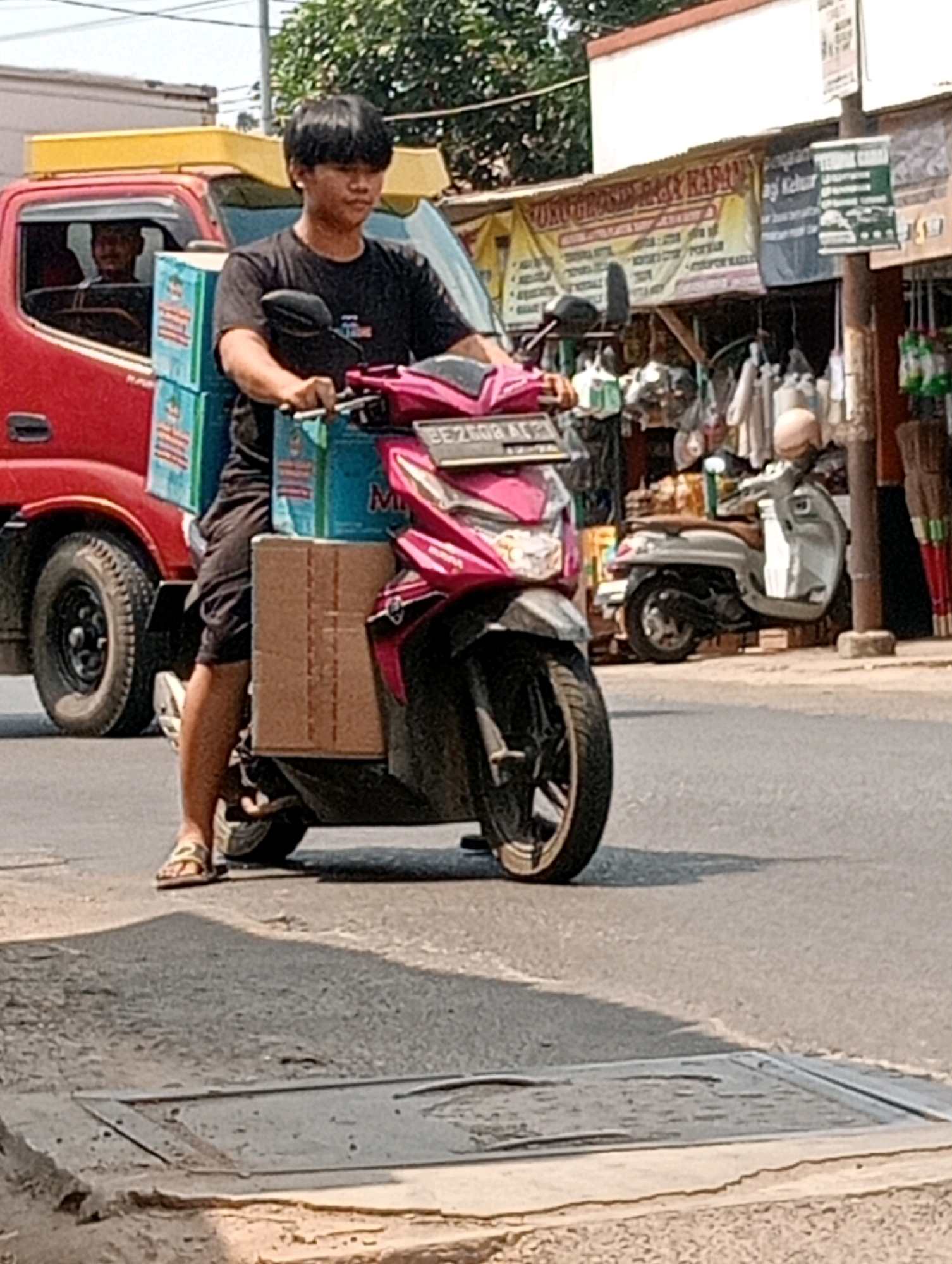 A young Indonesian male wearing a black t-shirt and black shorts rides a pink and black scooter on a paved street. He is carrying two stacks of blue and white boxes and a brown cardboard box on the scooter. In the background, a red truck is parked next to a market stall with various goods displayed. Another scooter, white and silver, is parked in front of the stall. The scene appears to be in a marketplace in Bandar Lampung, Indonesia, based on the presence of the truck, market stalls, and scooters typical of Indonesian street scenes. The time of day is likely daytime due to the bright sunlight and shadows cast on the ground.