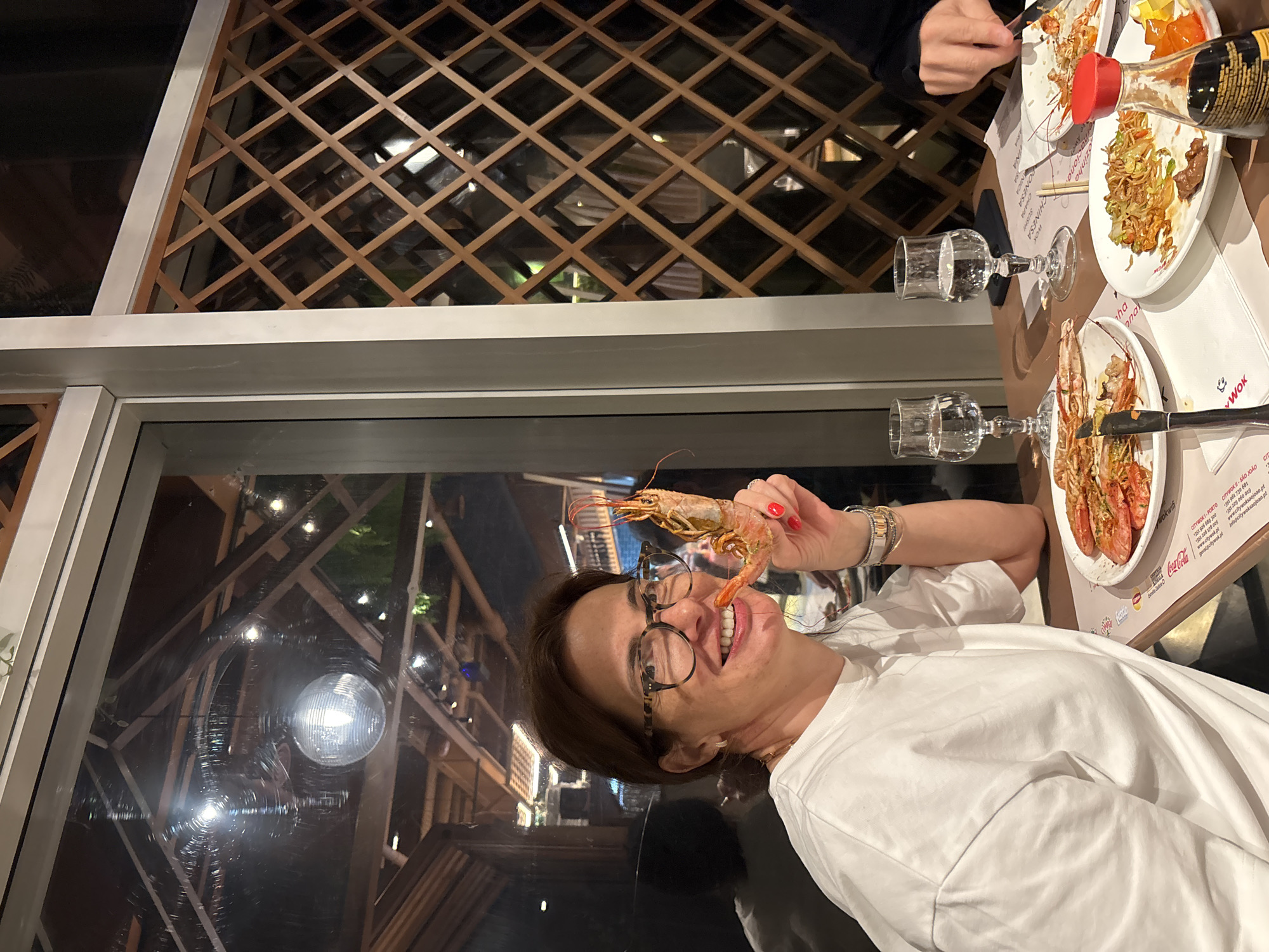 The image shows a woman in an indoor restaurant setting in Vila Nova de Gaia, Portugal, as suggested by text on the table menu. The woman, wearing glasses and a white t-shirt, is holding a cooked shrimp near her mouth and smiling. On the table are plates with food, including noodles, a bottle of soy sauce, cutlery, and glasses. The interior of the restaurant has a grid-like wooden design. Outside the window, lights and a decorative wooden structure are visible.