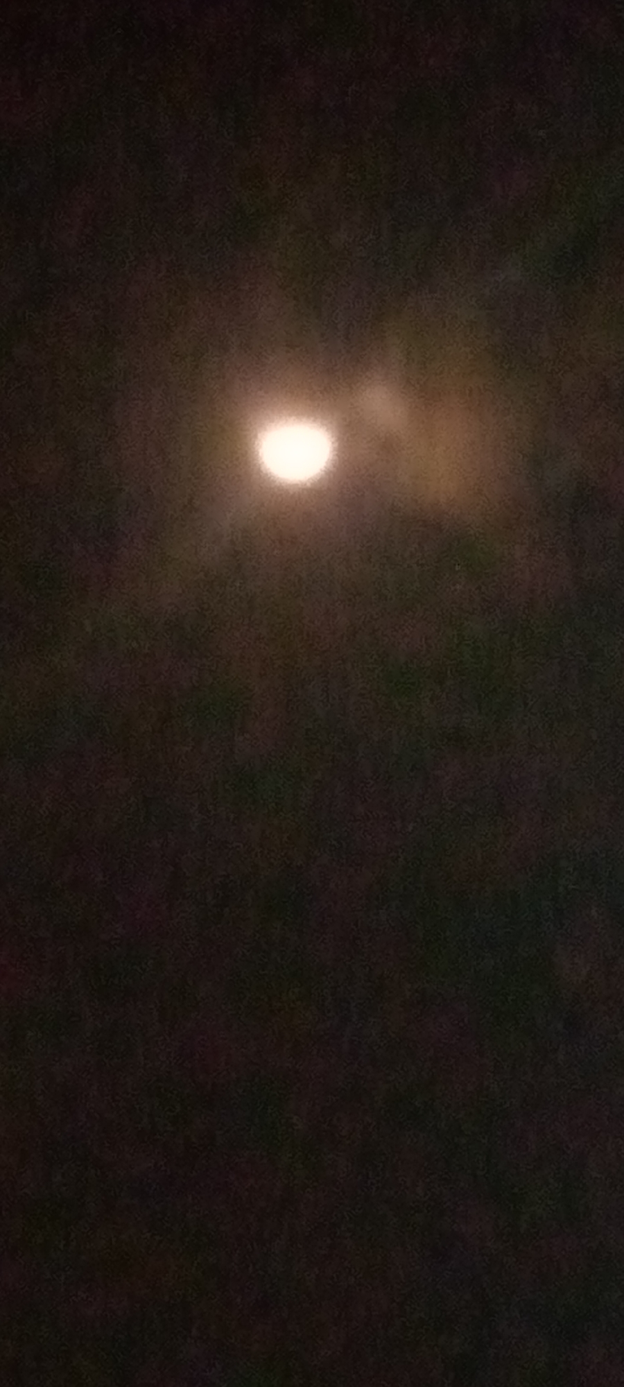 The image is a dark, out-of-focus photograph of the night sky. A bright, circular object, likely the moon, is visible in the upper center of the frame, surrounded by hazy clouds. The rest of the image is a deep, dark blur, suggesting a very low-light environment. There is no discernible information about the city of Sumberwono, Indonesia, or any specific details regarding people, subjects, objects, or actions.