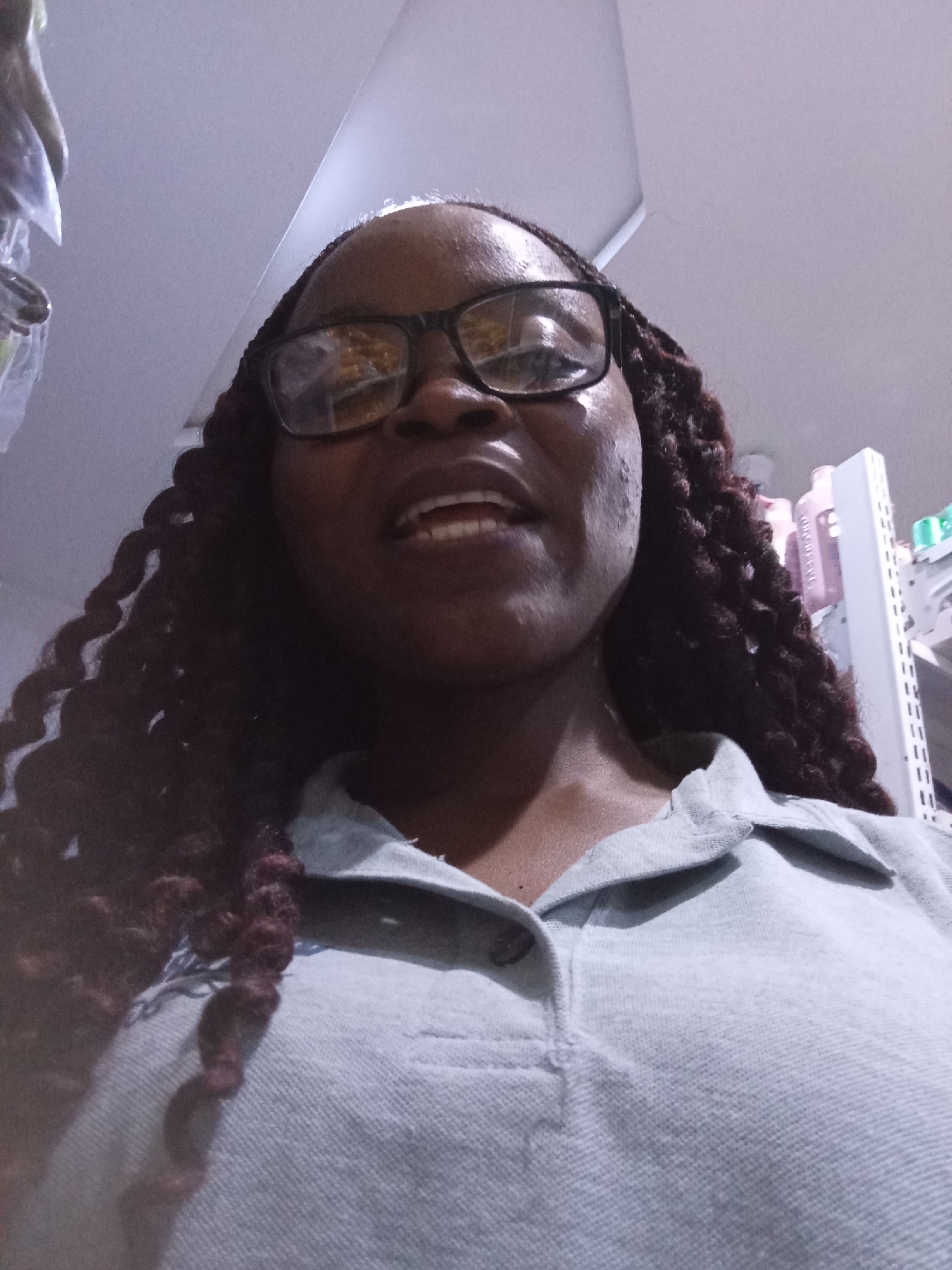 The media file shows a close-up, low-angle selfie of a Black woman with dark skin, wearing glasses and a light blue collared shirt. Her hair is dark, braided, and reaches down her shoulders. She appears to be smiling or laughing, with her mouth open and teeth visible. The background is blurred and shows shelves with various products, suggesting she might be in a store or a similar retail environment. The lighting is somewhat dim, and the overall impression is casual and candid. There is no discernible text visible in the image. The setting appears to be indoors, possibly a shop in Howayi, Nigeria, given the provided location context. The time of day and weather are not evident.