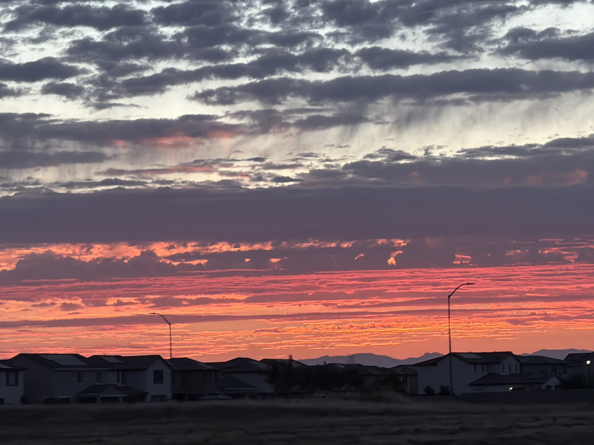 The image shows a dramatic sunset over a suburban neighborhood in Roseville, United States of America. The sky is filled with vibrant streaks of orange, pink, and purple clouds, with darker clouds in the upper portion of the frame. In the foreground, the silhouettes of houses and streetlights are visible against the colorful sky. The landscape appears to be dry and arid, with mountains visible in the distance. The overall impression is one of a breathtaking natural phenomenon occurring over a typical residential area.