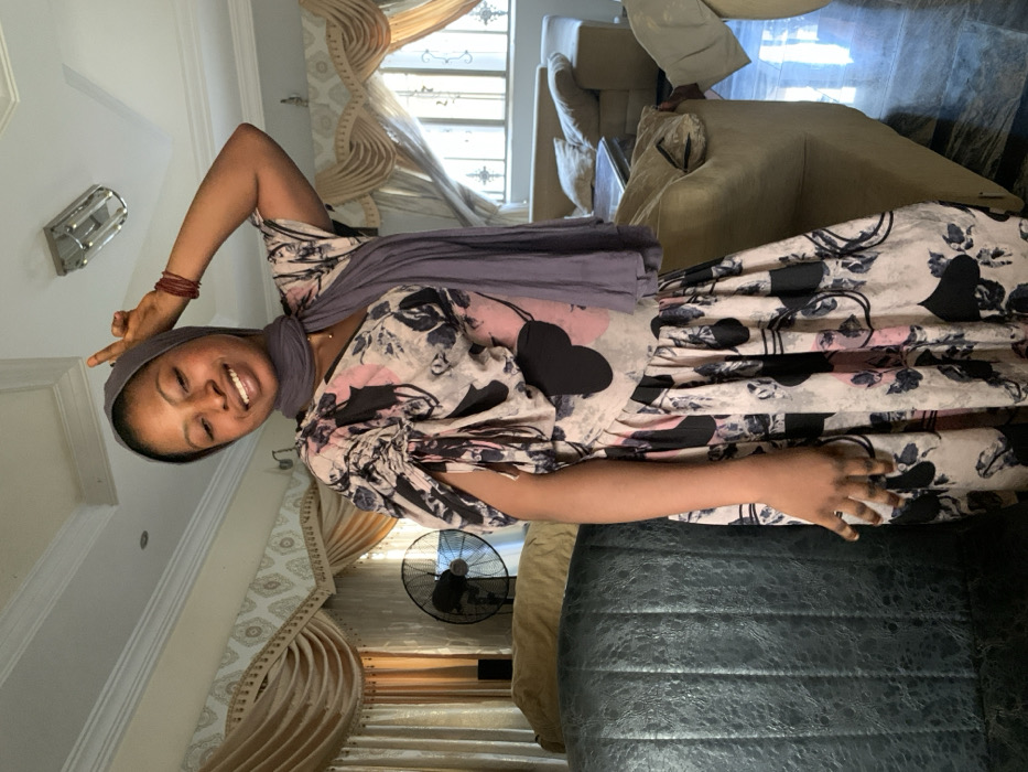 The image shows a smiling woman posing indoors, likely in a living room setting. The woman, who has dark skin, is positioned slightly to the left of the frame, facing the viewer with a broad, cheerful smile that shows her teeth. She is wearing a light-colored dress with a distinctive pattern featuring black hearts, pink accents, and black or grey floral motifs. A solid grey or light purple head covering is wrapped around her head and neck. On her right wrist, she wears several thin, reddish-brown bracelets. Her right arm is raised, with her hand forming a peace sign (V-shape with index and middle fingers), while her left hand rests casually near her waist with fingers slightly curled.
The background reveals a home interior. To the left, a silver or metallic rectangular light fixture with a slatted design is visible on the ceiling. Light-colored walls, possibly white or off-white, frame the scene. Behind the woman, decorative light-colored curtains, possibly cream or beige, with valances and drapes, are visible, suggesting a window letting in natural daylight. A black standing fan is clearly visible in the mid-ground to the right of the woman. In the foreground on the right, a dark-colored, possibly leather or faux-leather, textured armchair or sofa is prominent. Further in the background, several lighter-colored upholstered sofas or armchairs are discernible, some appearing to be recliners. The overall impression is one of a relaxed and happy moment captured in a brightly lit indoor space during the daytime.