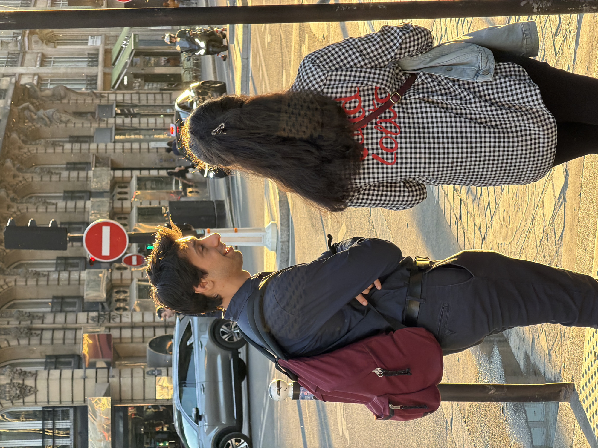 This image, oriented on its side, captures two individuals on a city street or sidewalk, bathed in the warm, golden light of late afternoon or early evening. In the foreground, a man with dark hair is seen looking upwards towards the left with a joyful expression, possibly laughing or smiling. He wears a dark blue jacket, dark pants, and a black belt, with a maroon backpack on his back. His arms are crossed.
Behind him, a woman is positioned with her back to the viewer. She has long dark hair, styled with a clip, and wears a black and white checkered shirt with the word "Love" embroidered in red script on the back. A light blue denim jacket is draped over her shoulder, and she wears black pants with a maroon strap visible, likely from a bag.
The urban background, consistent with Paris, France, features multi-story buildings with numerous windows and architectural details. A red circular "no entry" traffic sign with a white bar, along with a traffic light, is visible above the man. Several vehicles, including a silver car and motorcycles, are in the background. A black metal railing runs across the foreground, separating the subjects from the street. The bright, clear weather is indicated by the strong sunlight and shadows.