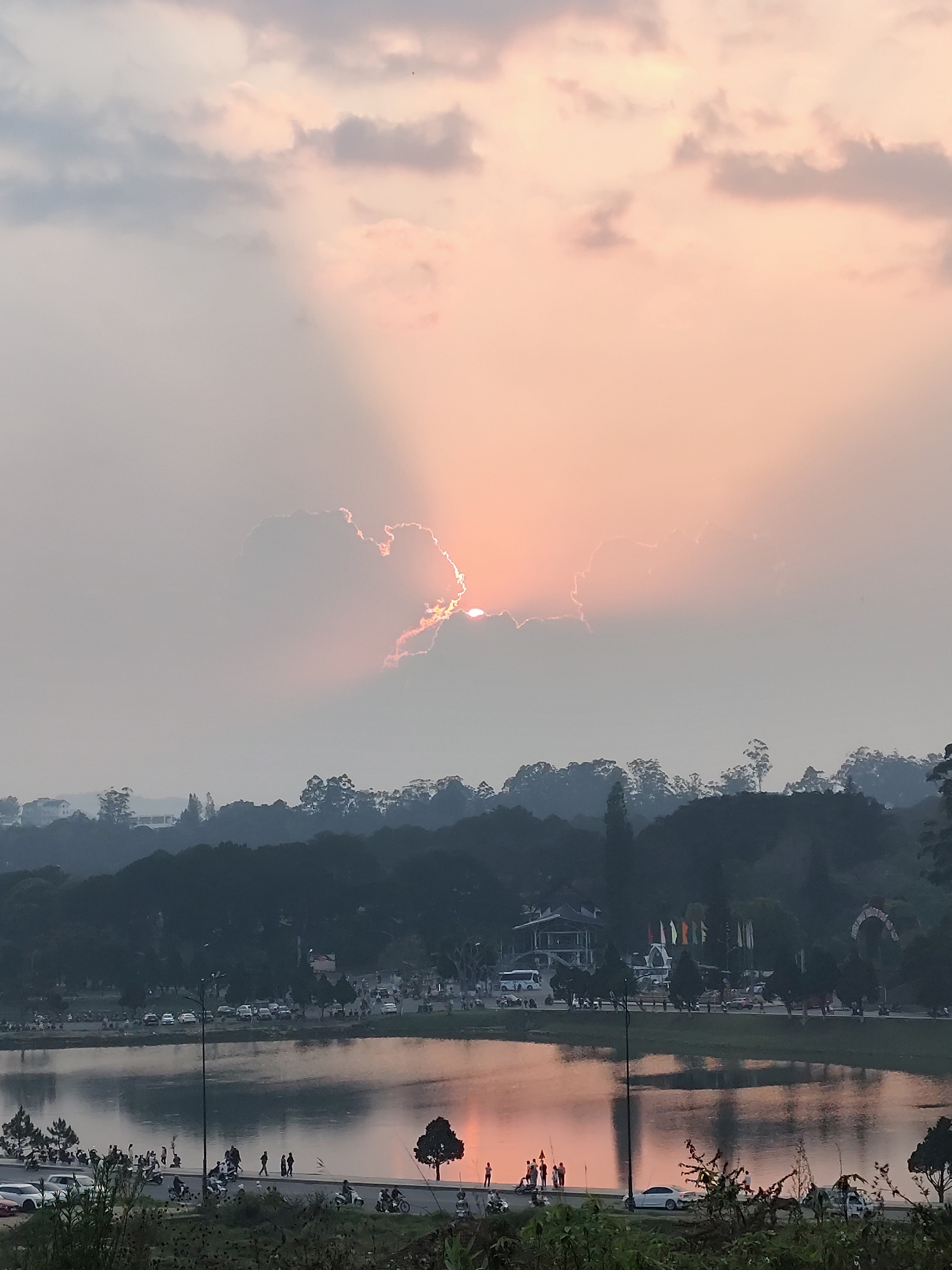 The image presents a panoramic view of Ho Xuan Huong Lake in Da Lat, Vietnam, during either sunrise or sunset. The sky exhibits a gradient of soft grey-blue in the upper left, transitioning to peach and orange tones towards the right, where the sun is partially obscured by a large cloud mass, emitting distinct crepuscular rays. The lake's surface accurately reflects the warm colors of the sky and the sun's illumination.
The far bank of the lake is characterized by a densely forested hillside, within which several light-colored buildings are discernible, along with a prominent, dark archway structure on the right. A paved road runs along the near shoreline, populated with numerous parked vehicles, including white cars and multiple motorbikes. Various individuals are present on the road and congregating in small groups near the water's edge, primarily on the left side of the frame, where they appear to observe the view. The immediate foreground is composed of dark, indistinct foliage.