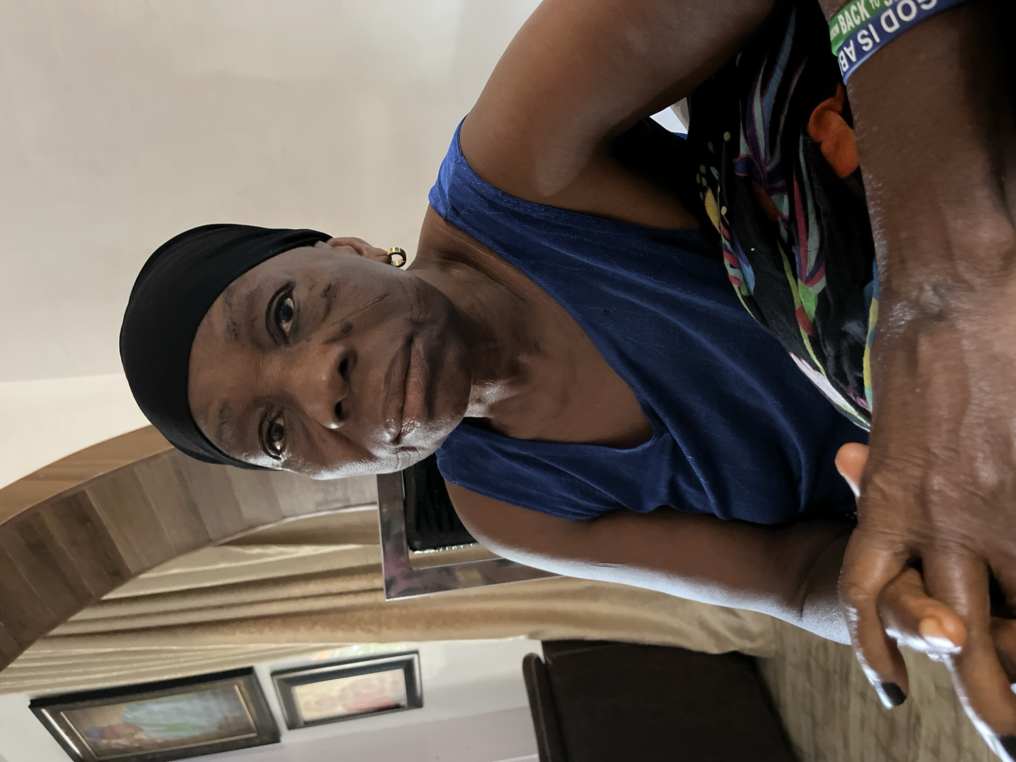 The image shows an older Black woman wearing a black headscarf and a blue tank top. She has visible facial scarring. A colorful patterned garment is draped around her shoulders. She is also wearing a bracelet that reads "GOD IS AB". Her hands are clasped together. The background shows a portion of a room with framed pictures on the wall and draped curtains. The location is likely a residential setting. The information about Jos, Nigeria is not verifiable from this image.