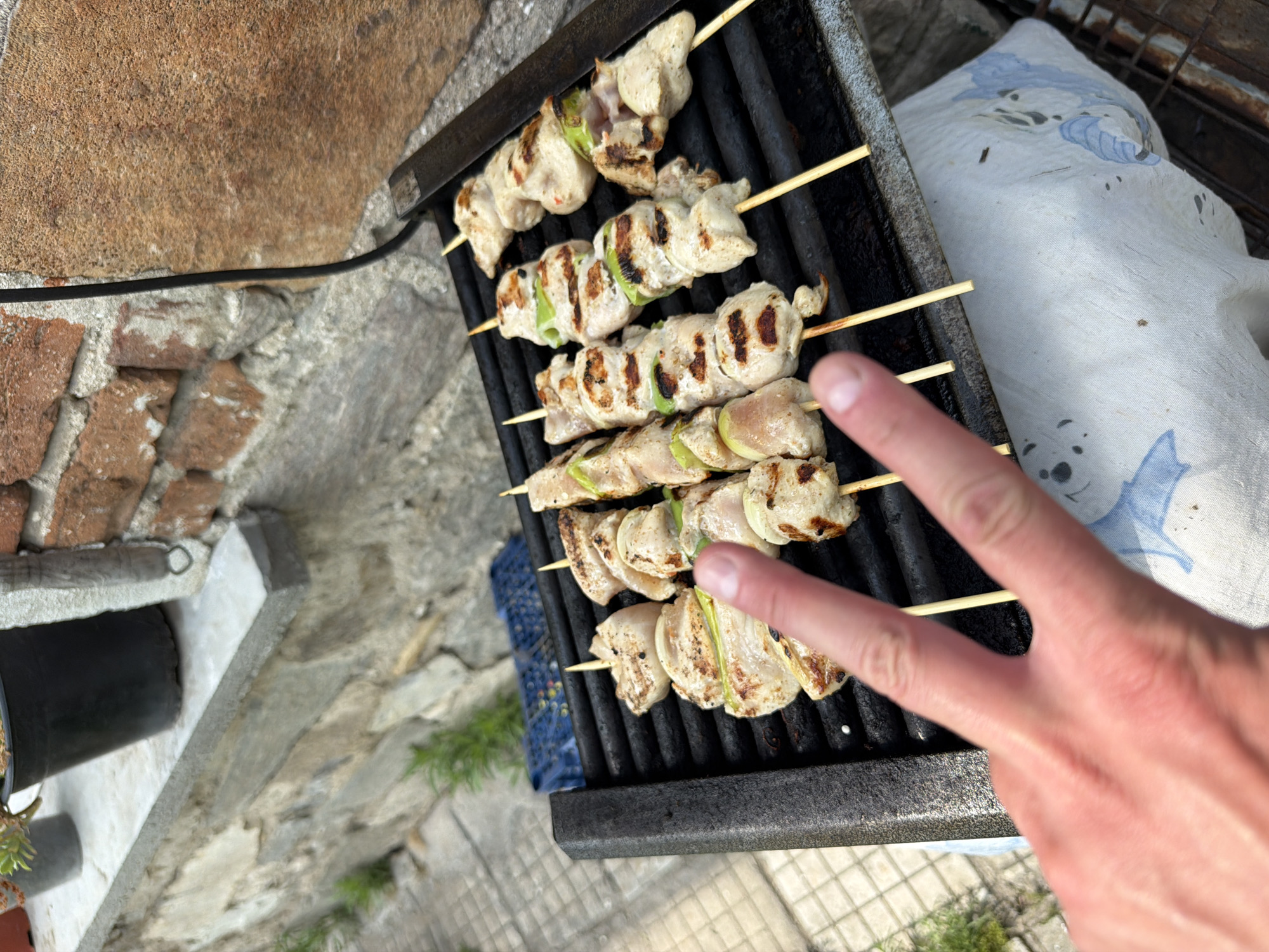 The image captures an outdoor grilling scene in Krumovo, Bulgaria. Six wooden skewers, laden with cubed chicken, onion pieces, and green bell pepper segments, are arranged on a black metal grill grate. The chicken displays visible char marks, indicating it is in the process of being cooked. A light-skinned human hand is positioned in the lower-left foreground, with the index and middle fingers extended in a V-shape, partially obscuring the grill. The background on the left consists of a low stone wall with a concrete cap, beyond which a tiled ground with sparse vegetation is visible, along with a partially obscured blue plastic crate. On the right, a reddish-brown brick wall and a darker, textured stone surface are present, intersected by a black electrical cable. A white cloth with light blue patterns is visible beneath the grill, serving as a covering for the surface below. Two out-of-focus potted plants are situated on the stone wall in the upper-left background.