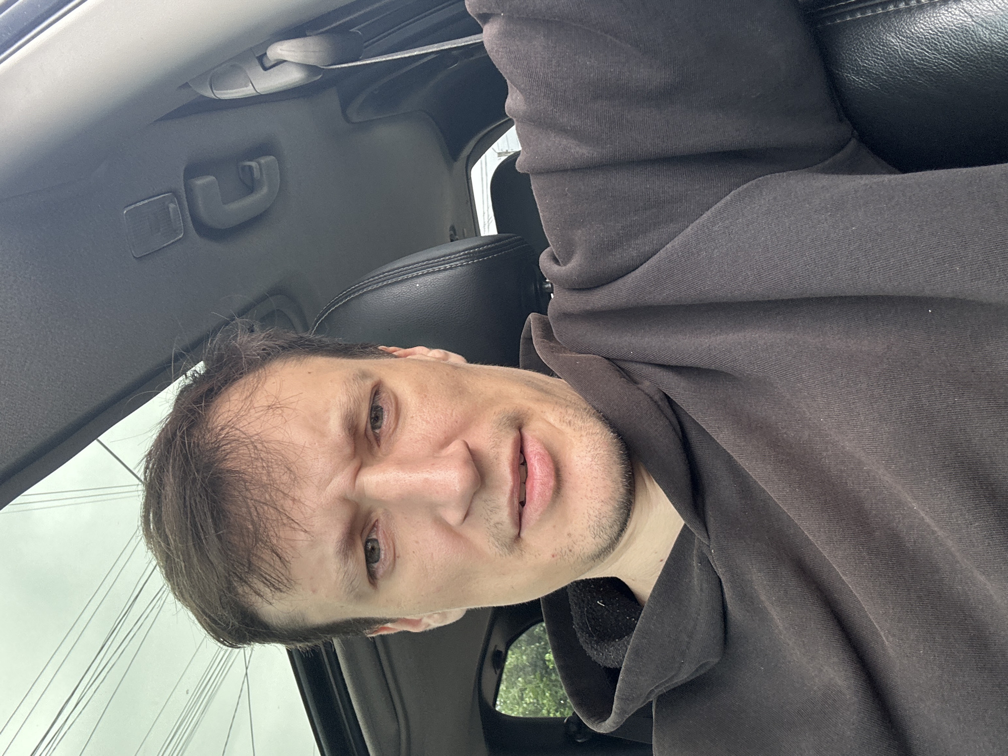 A close-up selfie shows a man in a dark hoodie inside a vehicle. He has brown hair, light skin, and is looking directly at the camera with a furrowed brow. The interior of the car is visible, including the headrest of a black leather seat, the roof lining with a grab handle, and part of the sunroof. The man's facial expression suggests concern or exertion. The exterior of the car is seen through the sunroof and a side window, showing a cloudy sky and some greenery. There are no other people or objects in the immediate foreground. The location is not specified in the image, nor is there any information about Makiv, Ukraine.
