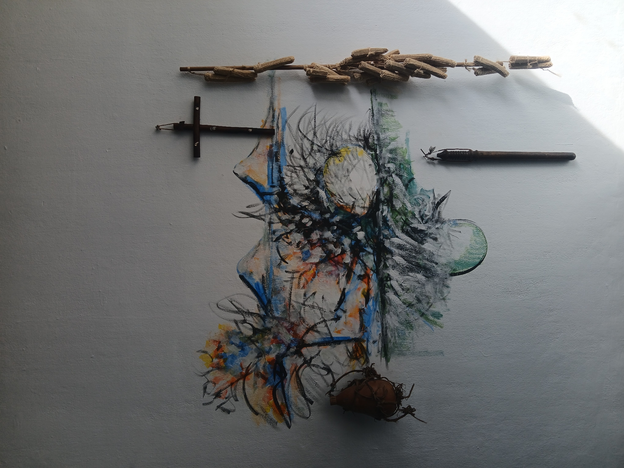 This is an image of an artistic installation on a light gray wall, likely indoors. At the top, a row of dried corn cobs is suspended on a twig or thin stick. Below, to the left, is a dark, wooden cross-shaped object. In the center is an abstract drawing/painting in various colors, including white, black, blue, orange, and green, resembling a burst of energy or an organic form. Below the painting sits a small, brown gourd-like object with attached, spiky tendrils. To the right of the cross, near the top, is a dark, vintage pen with its cap attached. The wall is unevenly lit, with brighter highlights in the upper right corner. There is no evidence or information in the image that would allow us to place this scene in Guayana City, Venezuela, or any other specific location.