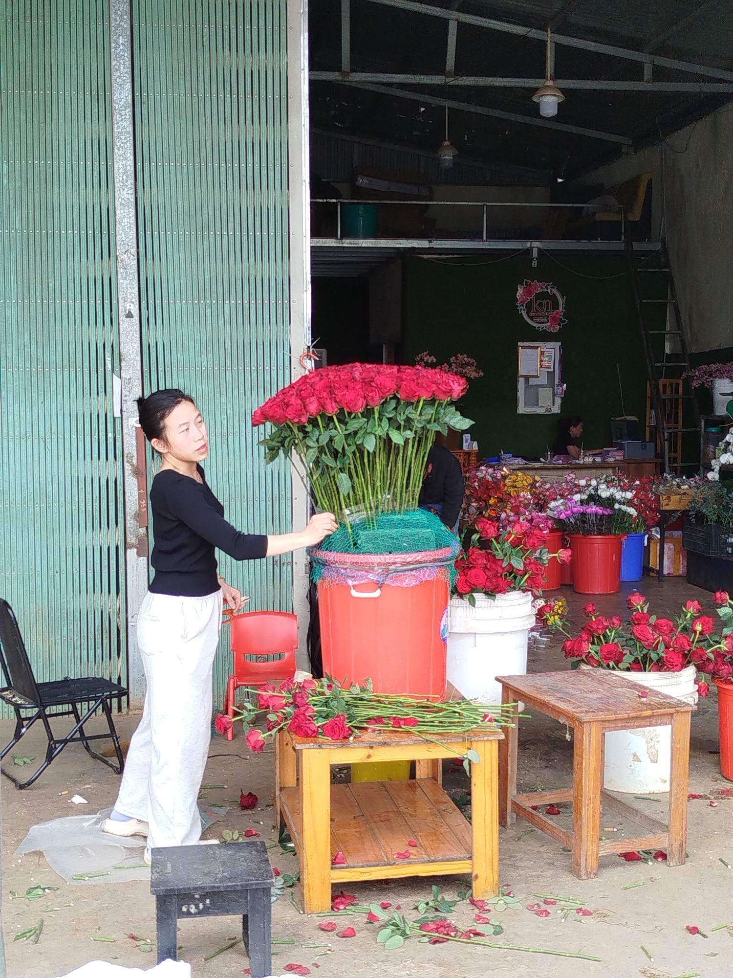 The image shows a young woman standing near a large arrangement of red roses at a flower shop in Cam Ly - Đà Lạt, Vietnam. She is wearing a black long-sleeved shirt and white pants. The roses are arranged in a green net atop a red bucket. A wooden table sits nearby, covered with loose roses and stems. Petals are scattered on the ground. The shop's open doors reveal a person sitting at a desk, more flowers, and a ladder leading to a mezzanine. The shop has a green wall with a floral wreath logo. A green corrugated metal door is partially open on the left. Chairs and stools are scattered around.