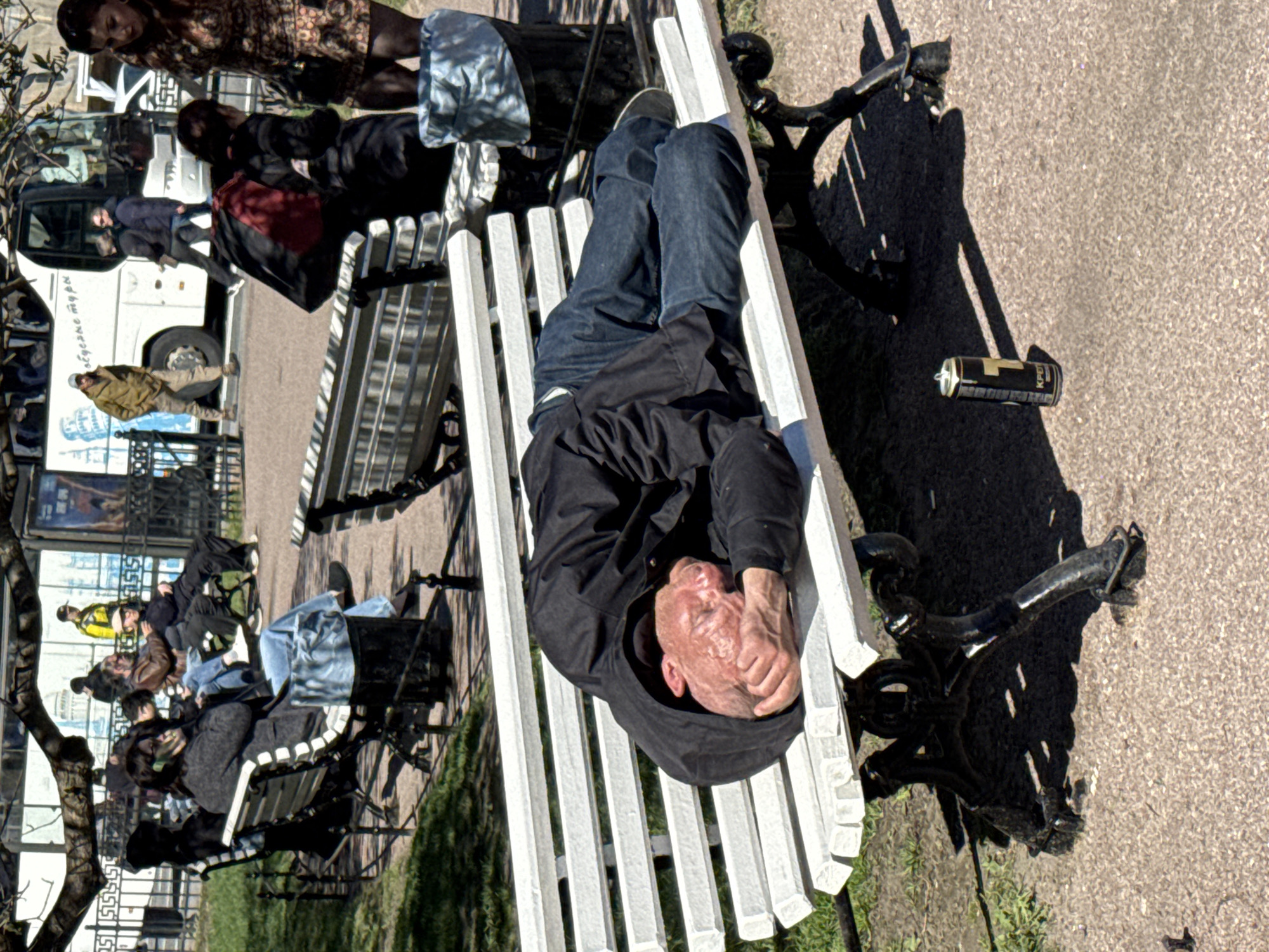 A man lies prone on a white park bench in Saint Petersburg, Russia. The man is wearing jeans and a black hooded top. His hands are clasped near his face. To the right of the bench is an empty can. In the background, other benches are occupied by people. A white tour bus is also visible. The ground is paved. The weather appears sunny and clear.